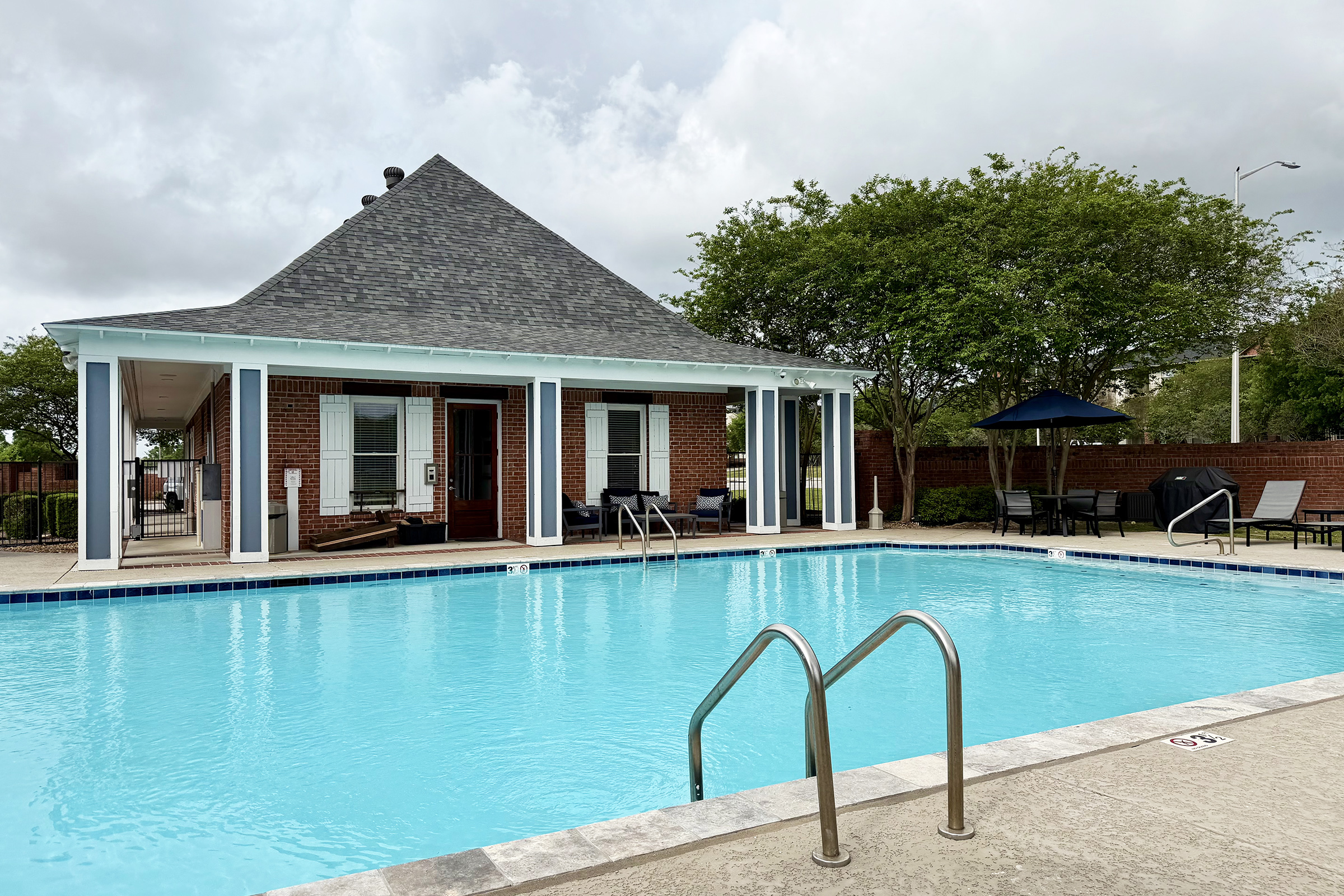 A clear blue swimming pool surrounded by a patio area featuring lounge chairs and tables. In the background, there is a brick building with large windows and a peaked roof, along with trees and a shaded umbrella nearby. The sky is partly cloudy.