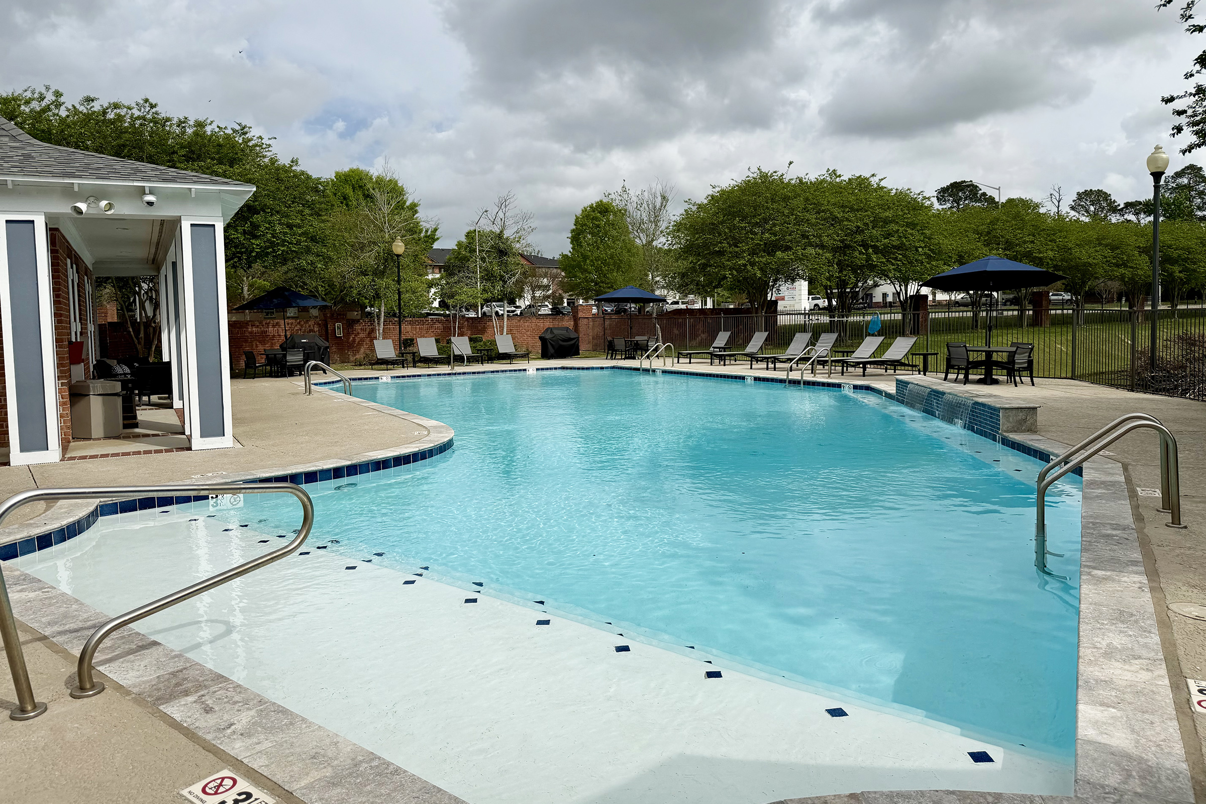 A calm outdoor swimming pool surrounded by lounge chairs and umbrellas. The water is clear and inviting, with a patio area to the side featuring a pool house. Lush greenery lines the background under a partly cloudy sky, creating a relaxing atmosphere.