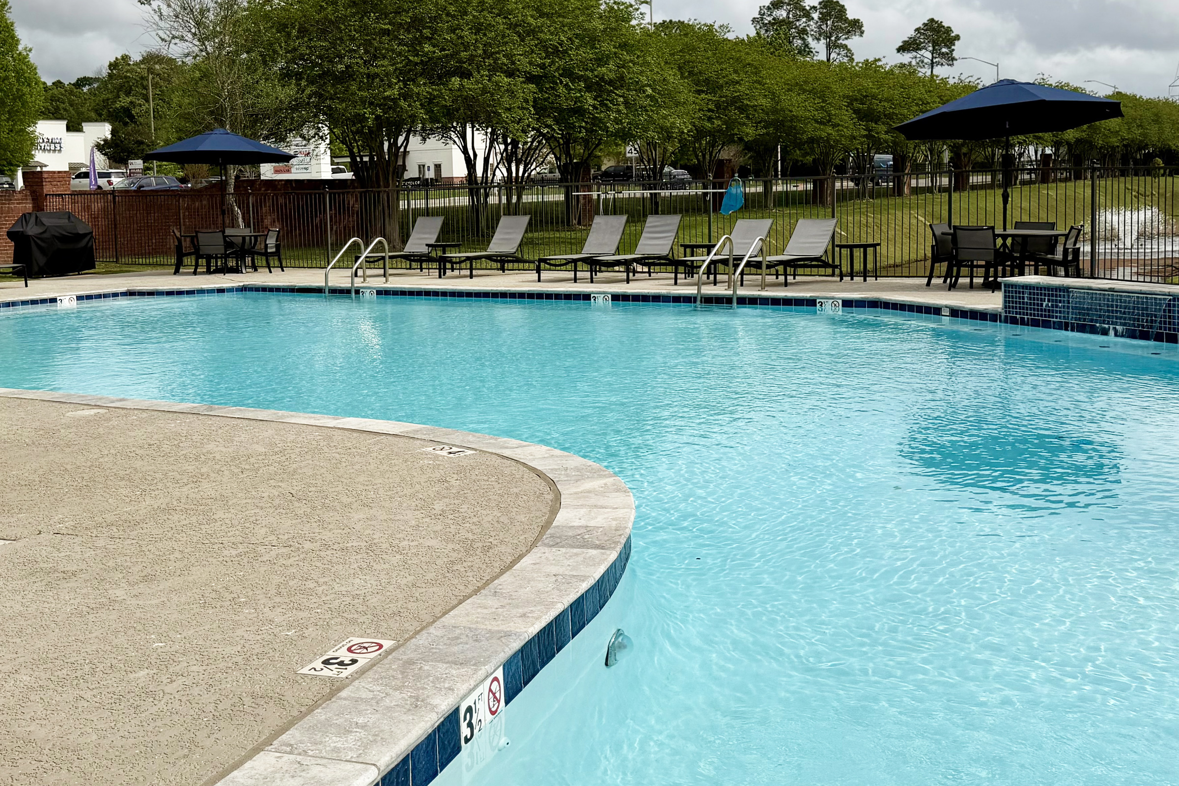 A clear blue swimming pool with a curved edge, surrounded by lounge chairs and umbrellas. In the background, there are grassy areas and trees. The pool area is well-maintained, with safety depth markers visible at the edge.