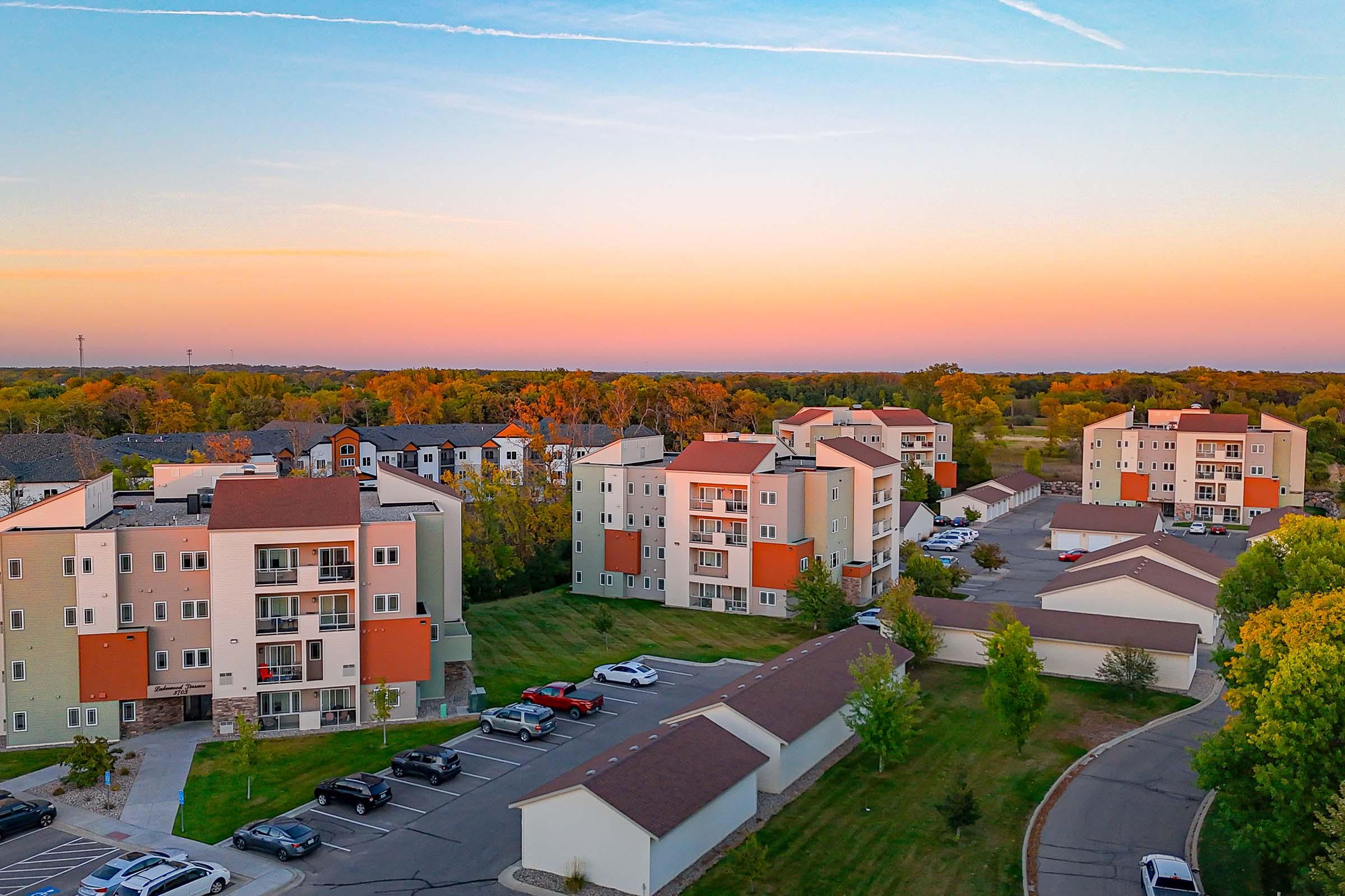Aerial view of a residential area featuring modern apartment buildings with varied colors. The scene includes green lawns, parked cars, and tree-lined surroundings, bathed in soft sunset hues of orange and pink.