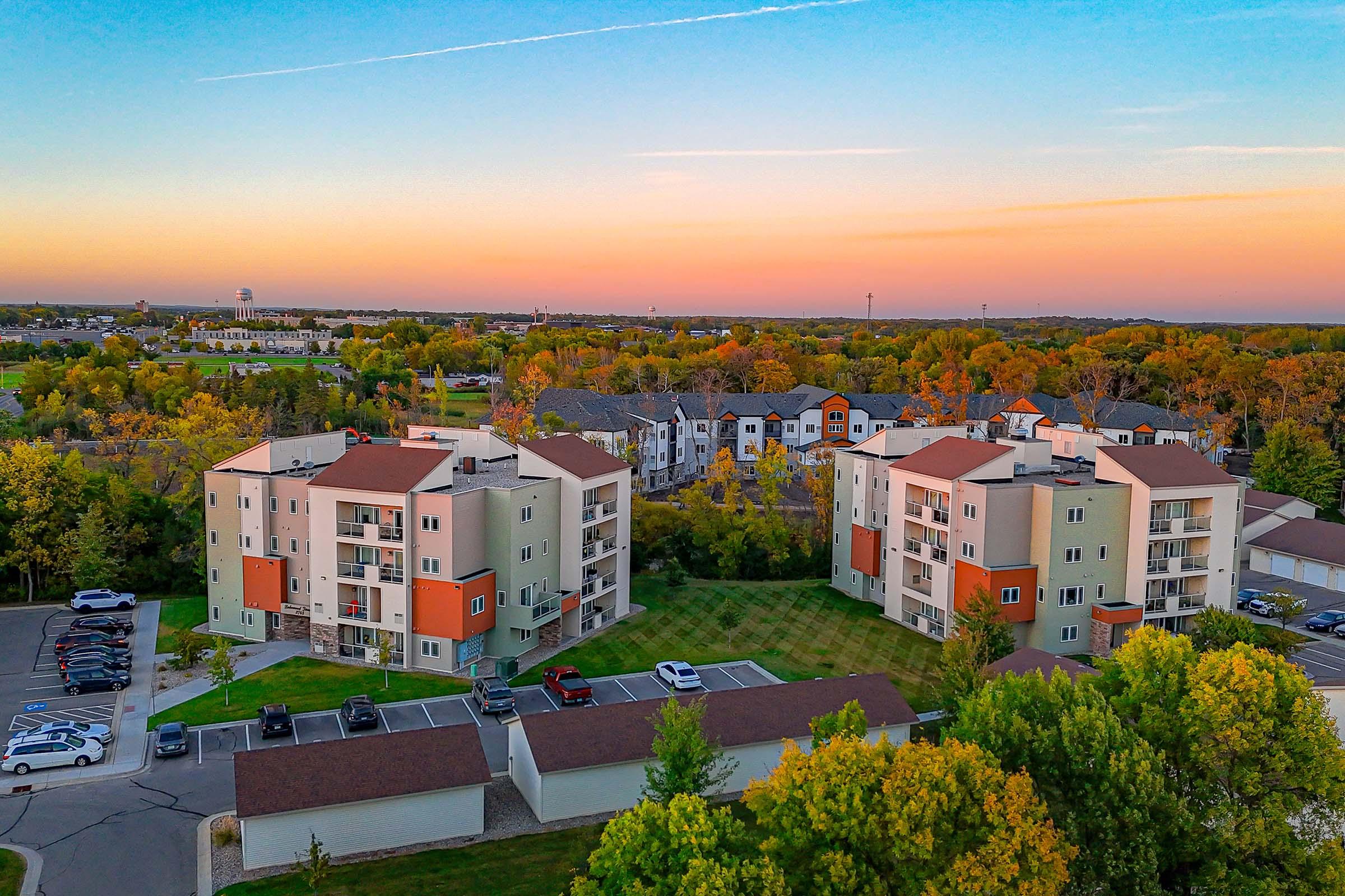 Aerial view of residential buildings with modern architecture, surrounded by greenery and trees displaying autumn colors. The sky features a gradient of sunset hues, and parking lots are visible in the foreground. The scene conveys a serene suburban atmosphere.
