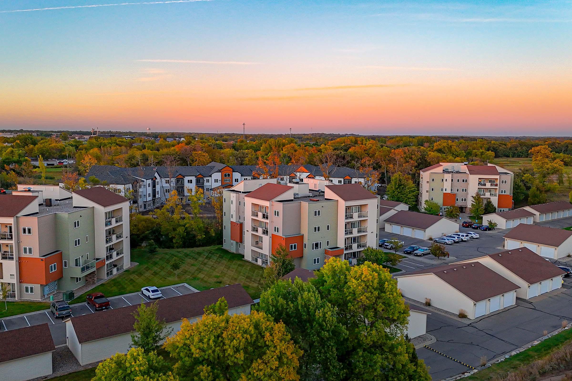 Aerial view of a suburban neighborhood featuring multiple residential buildings and garages surrounded by greenery. The sky transitions from blue to warm hues of orange and pink during sunset, highlighting the peaceful atmosphere of the area.
