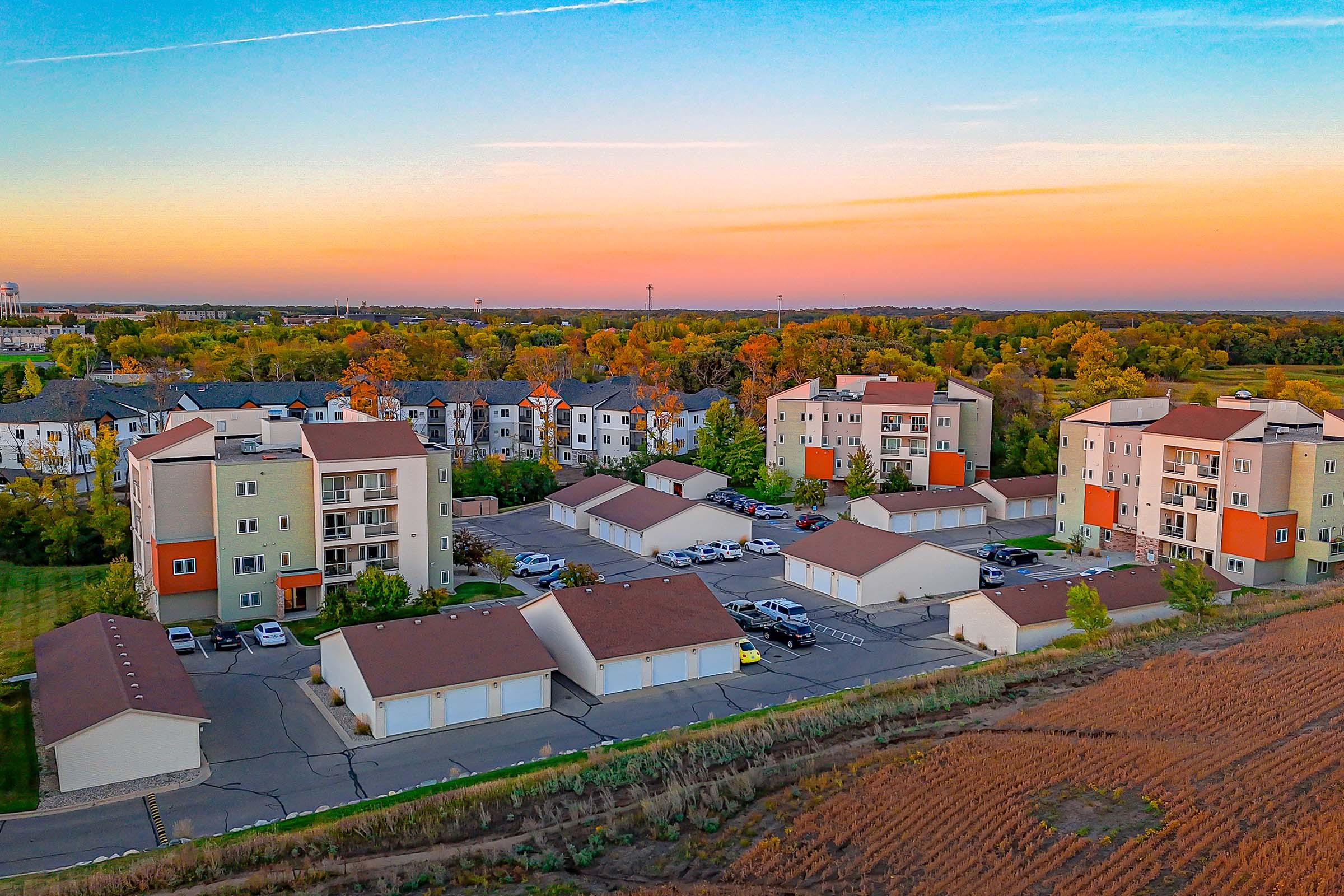 Aerial view of a residential area featuring several multi-story apartment buildings and a cluster of garages. The landscape includes trees with autumn foliage and a sunset sky, creating a vibrant atmosphere. Nearby, a field is visible, adding to the suburban setting.