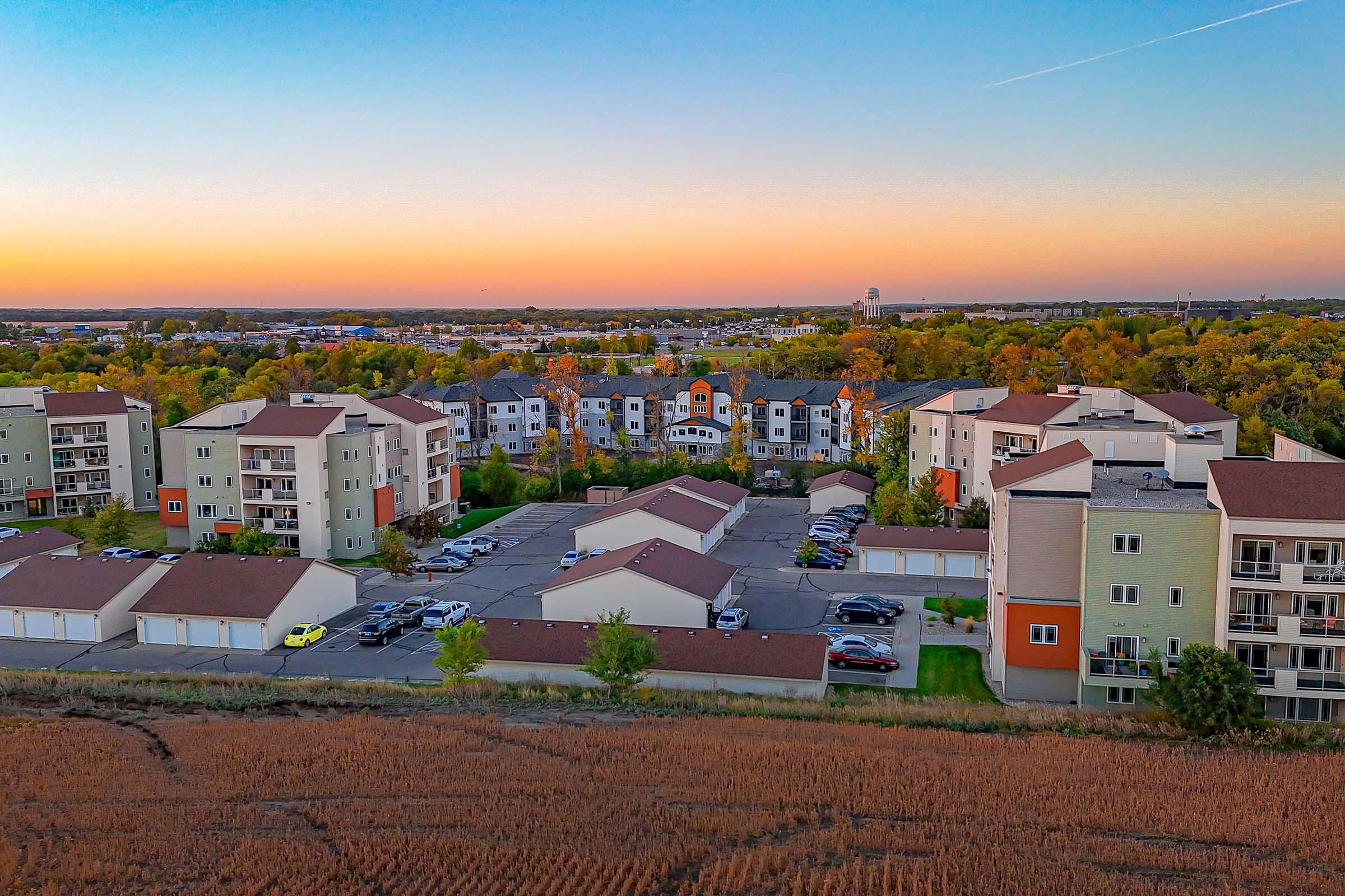 Aerial view of a residential area featuring modern apartment buildings with varying colors and designs. The foreground includes a field, and in the background, trees display autumn colors under a clear sky at sunset, creating a warm, inviting atmosphere. Parking lots and green spaces are visible throughout the neighborhood.