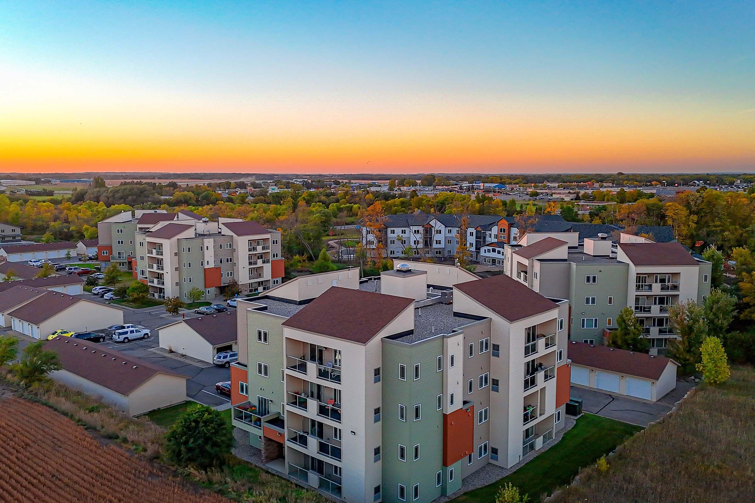 Aerial view of residential apartment buildings with varying colors, surrounded by greenery and open fields. The horizon showcases a sunset with vibrant orange and blue hues, creating a peaceful atmosphere. Cars are parked nearby, and additional housing can be seen in the background.
