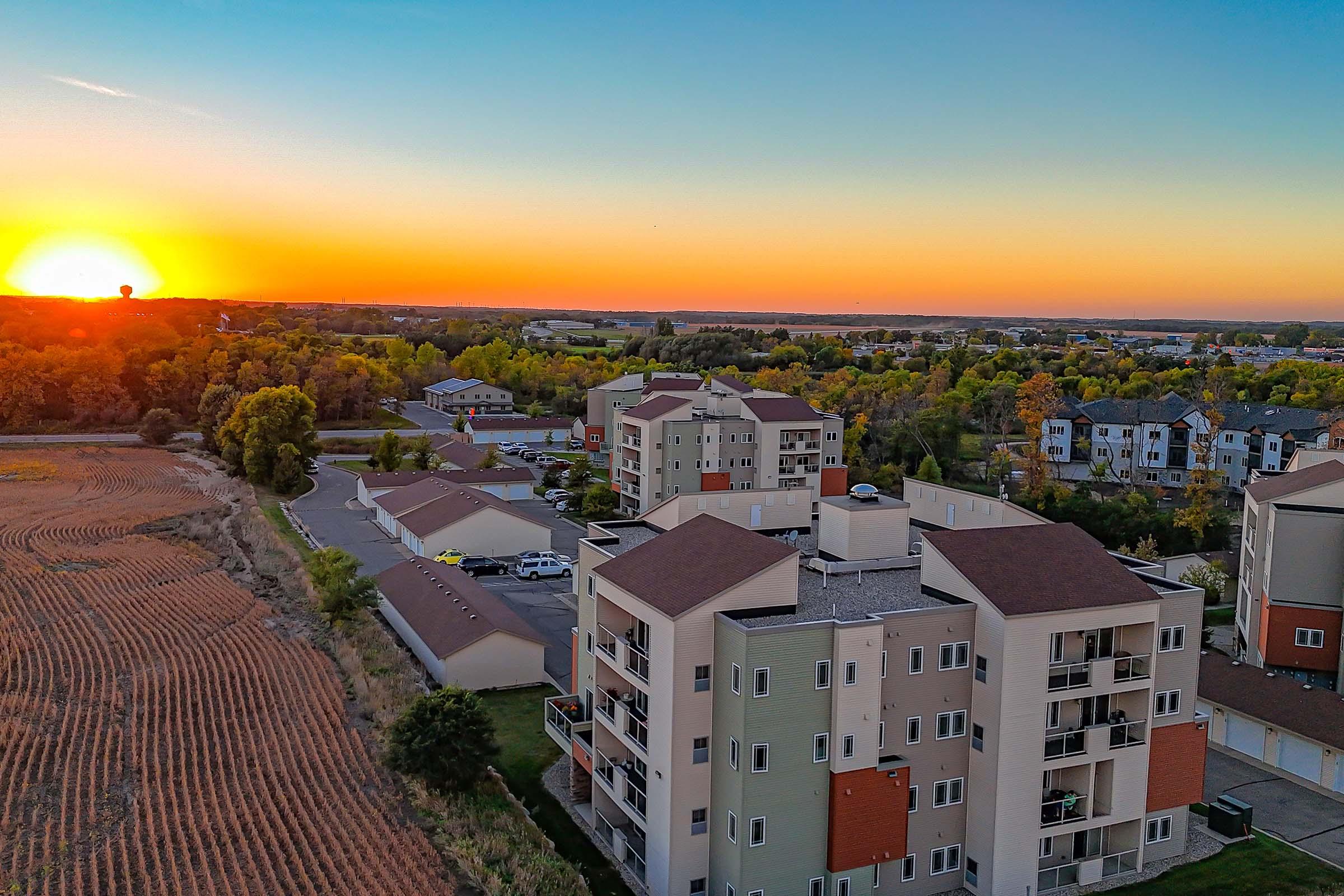 Aerial view of a residential area at sunset, showcasing several multi-story buildings and a backdrop of trees and fields. The sky displays vibrant orange and yellow hues as the sun sets on the horizon, creating a tranquil evening atmosphere.