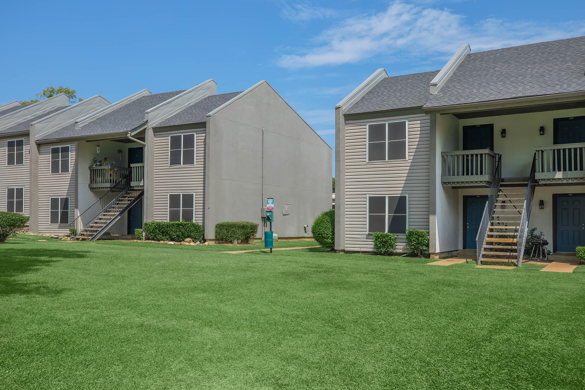 Two multi-family residential buildings with light gray and beige siding. Each building features stairs leading to the second floor units, surrounded by manicured green grass and small shrubs. A clear blue sky and a few clouds can be seen in the background.