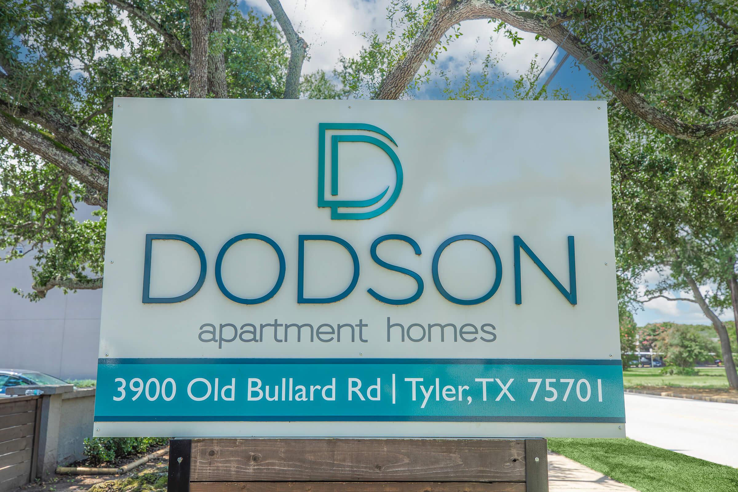 Sign for Dodson Apartment Homes, featuring the logo and the address: 3900 Old Bullard Rd, Tyler, TX 75701. Surrounded by trees and blue sky, the sign is modern and clear, providing essential information for potential residents.