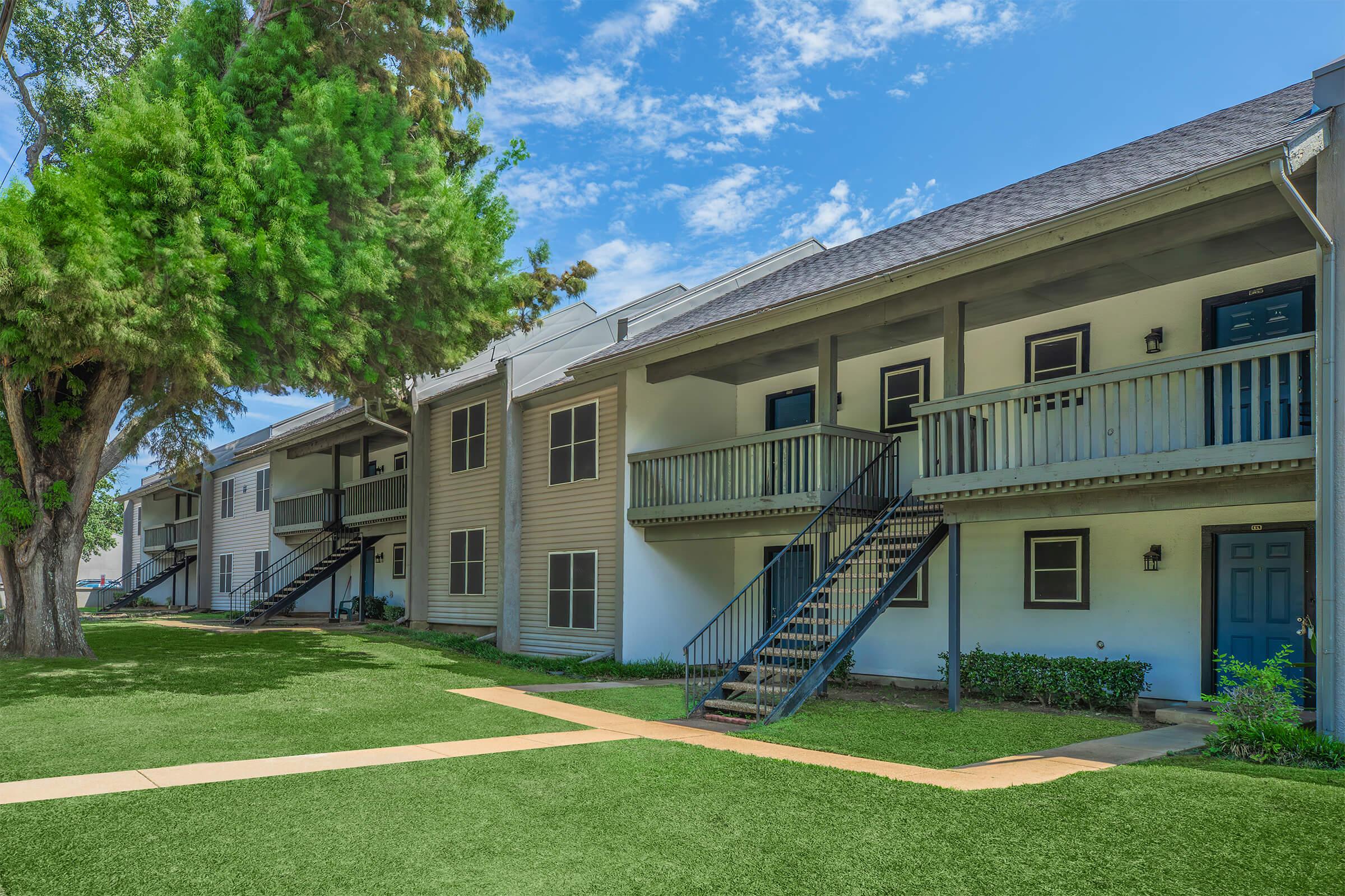 A well-maintained apartment complex featuring two-story buildings with green lawns and trees. The architecture includes balconies and stairs leading to individual units, under a bright blue sky with scattered clouds.