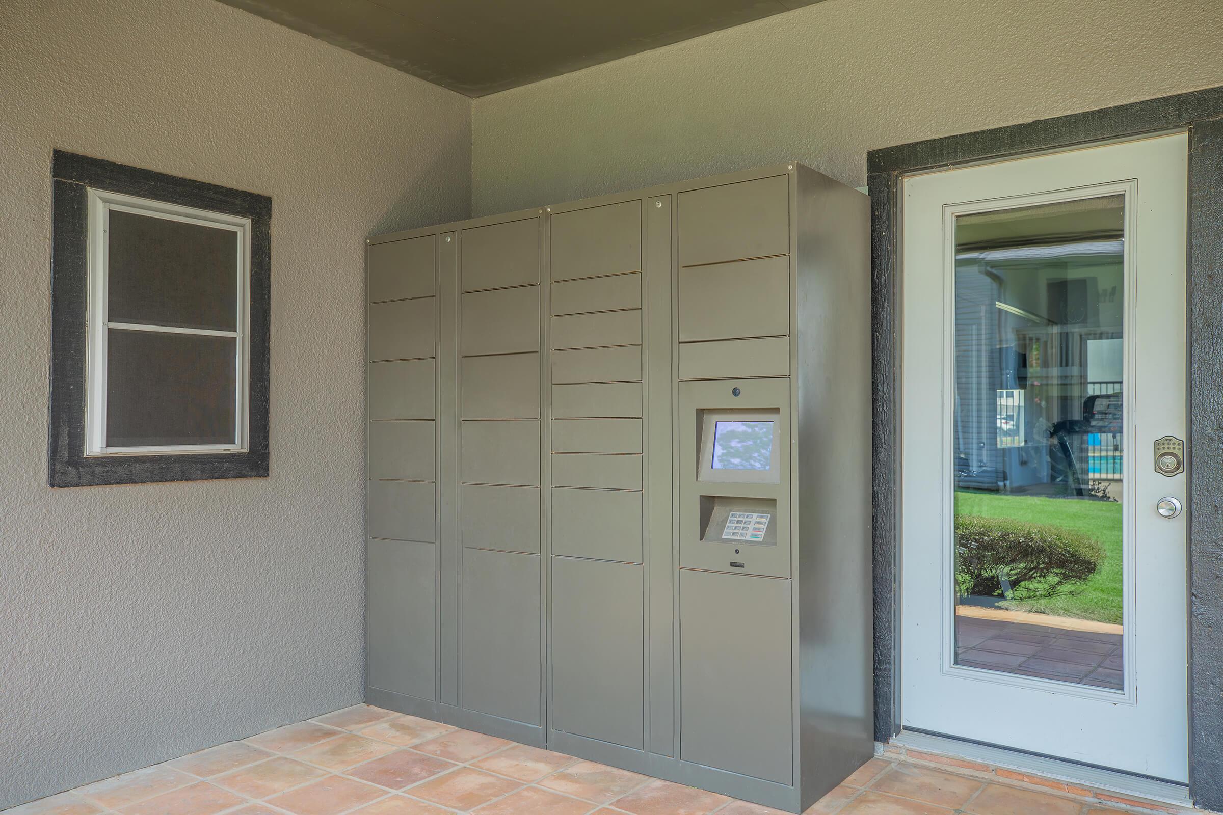 Delivery locker storage unit installed next to a door. The unit has multiple sections for packages, a keypad interface, and is situated on a tiled floor. A window is visible to the left of the locker, and the door on the right leads outside to a lawn area.