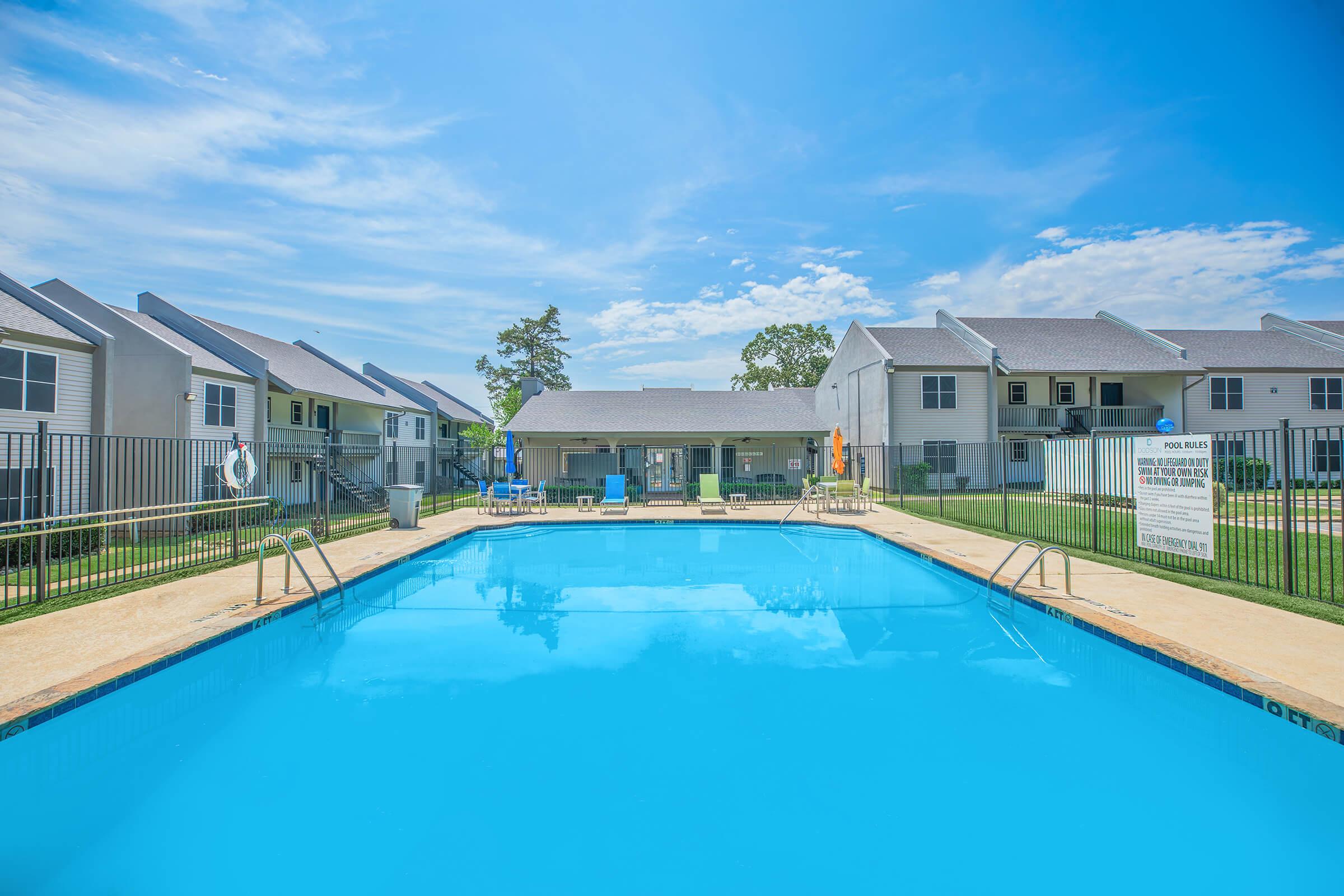 A clear blue swimming pool surrounded by a well-maintained lawn. Apartment buildings are located in the background, with a clubhouse visible near the pool area. The sky is bright with scattered clouds, creating a pleasant summer atmosphere.