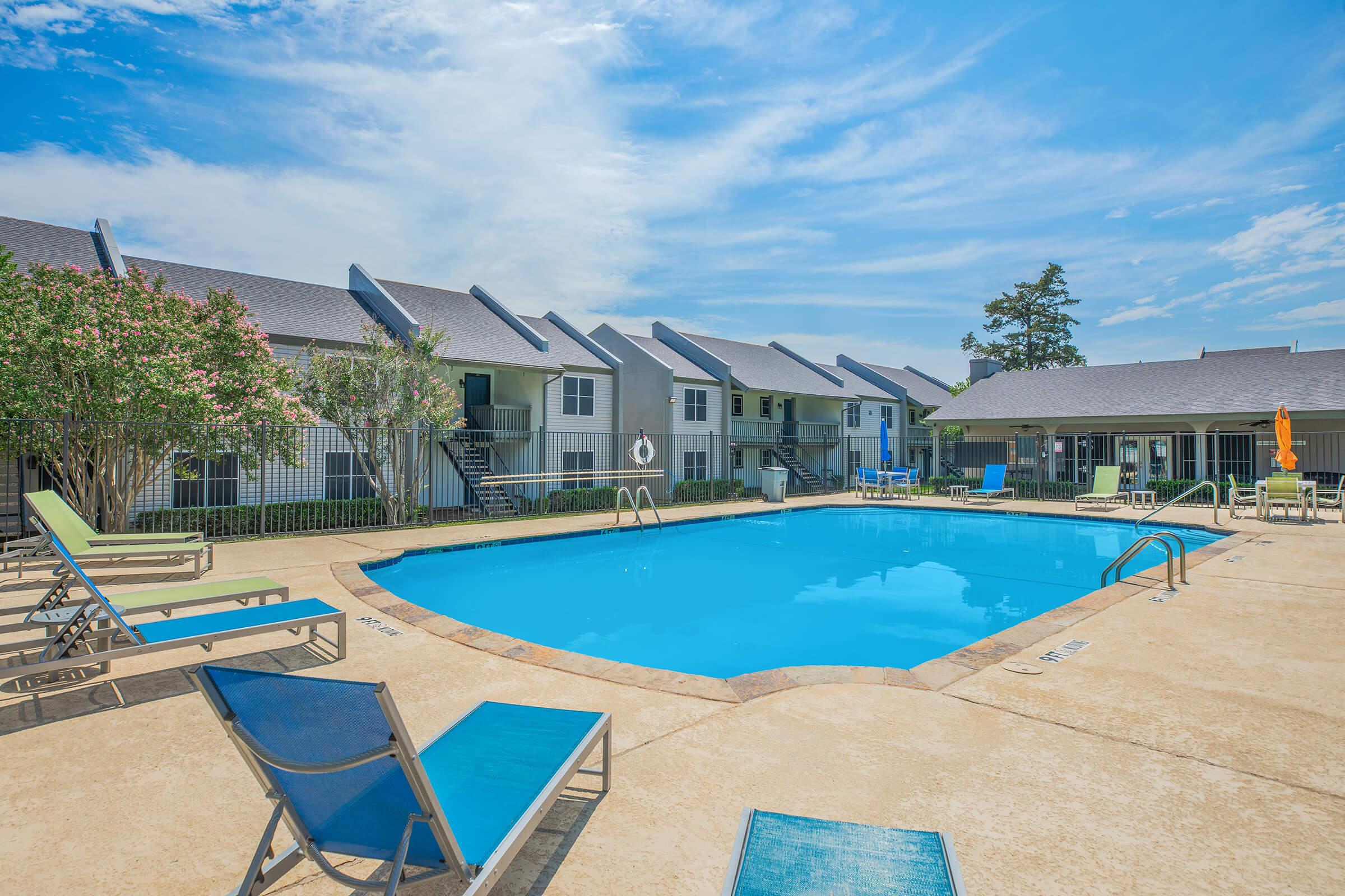 A sunny pool area features a clear blue swimming pool surrounded by lounge chairs. In the background, there are multiple two-story buildings with balconies, greenery, and an outdoor umbrella for shade, creating a relaxing atmosphere ideal for leisure and sunbathing.