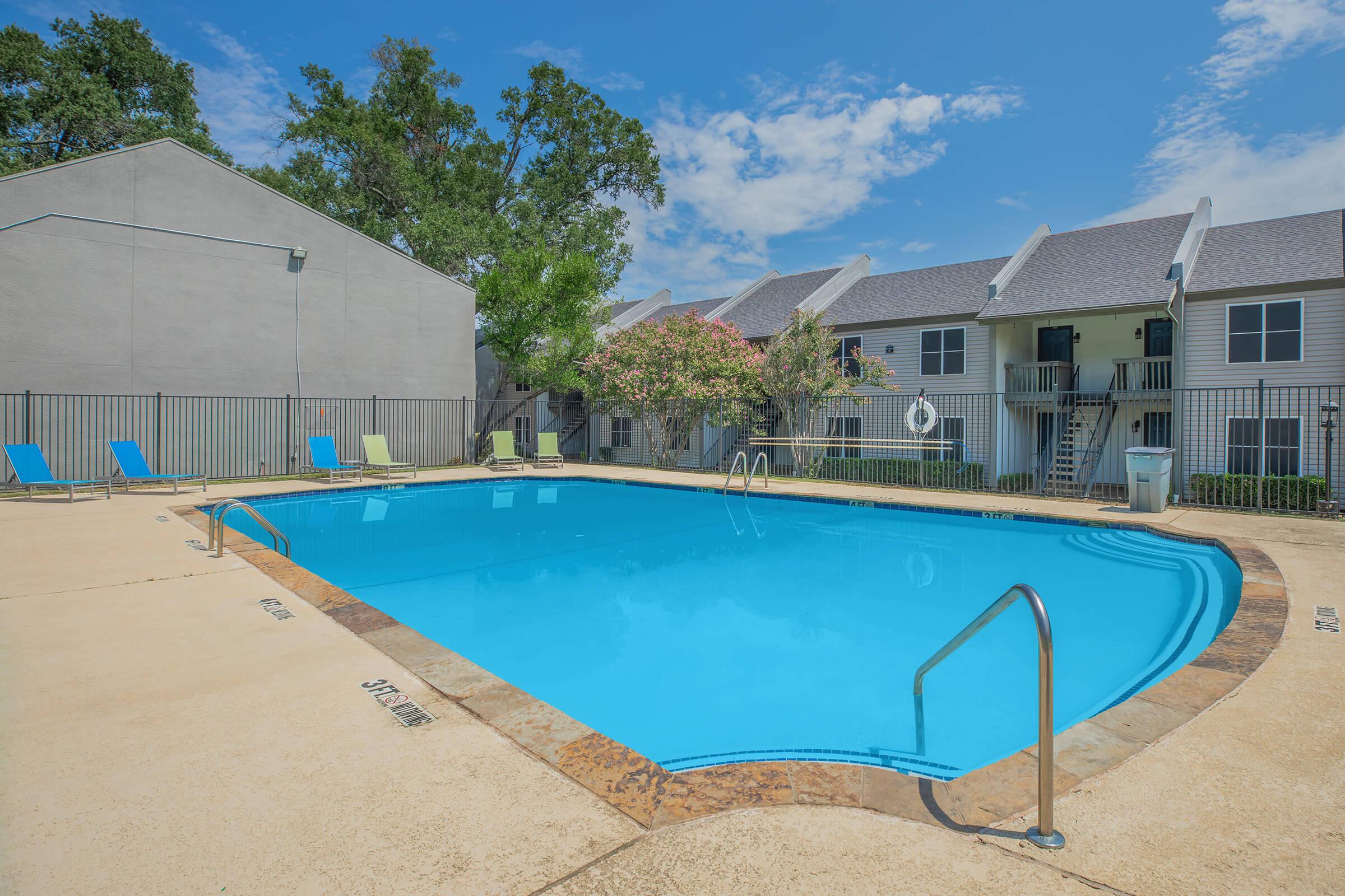 A clear blue swimming pool surrounded by a tan deck, with lounge chairs arranged nearby. In the background, two-story apartment buildings are visible, partially shaded by trees under a bright blue sky with a few clouds. The pool area appears inviting and well-maintained.