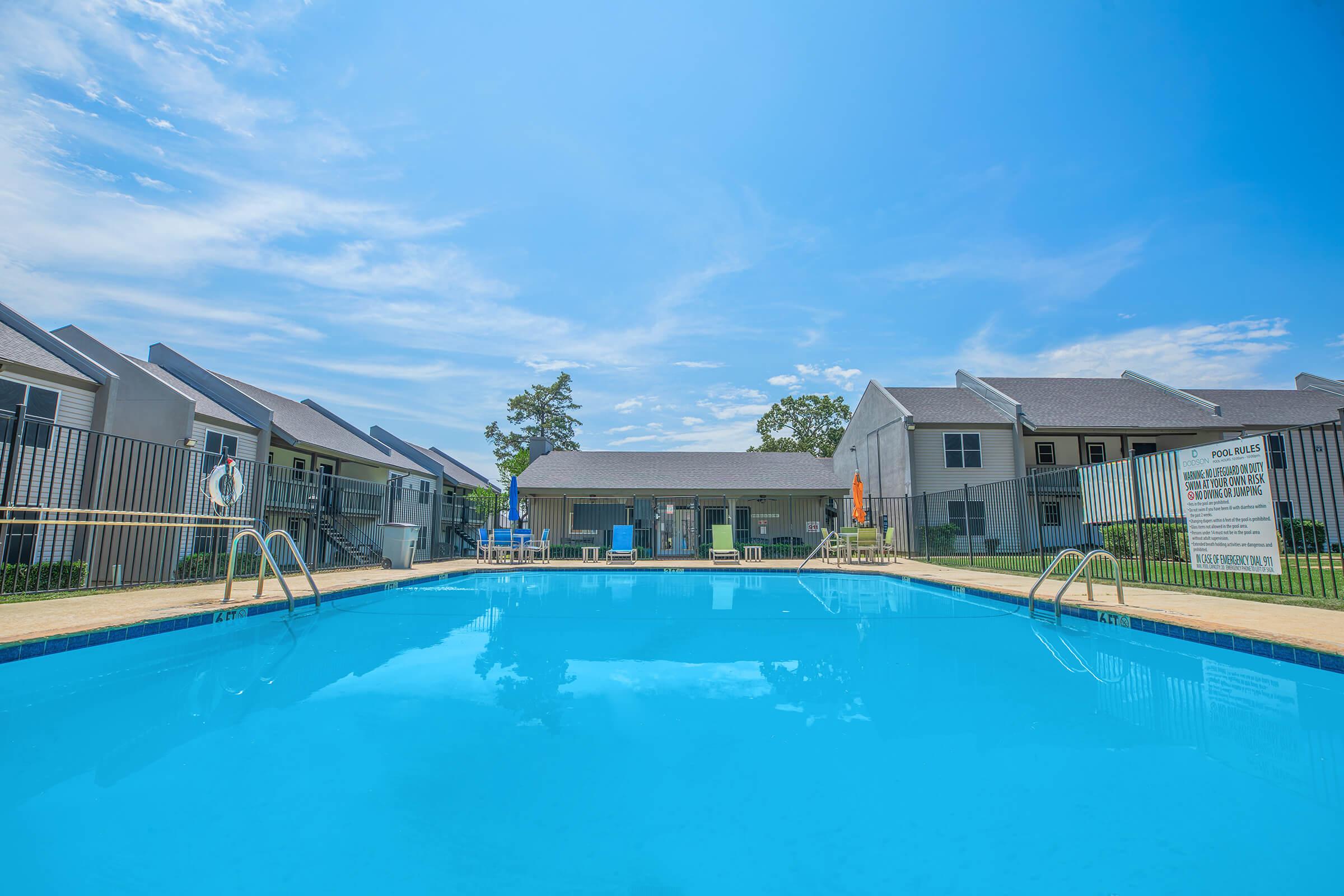 A bright blue swimming pool surrounded by a grassy area and residential buildings. There are lounge chairs and umbrellas by the poolside, with a clear blue sky overhead and a few clouds. The area appears inviting and well-maintained, suitable for relaxation and leisure activities.
