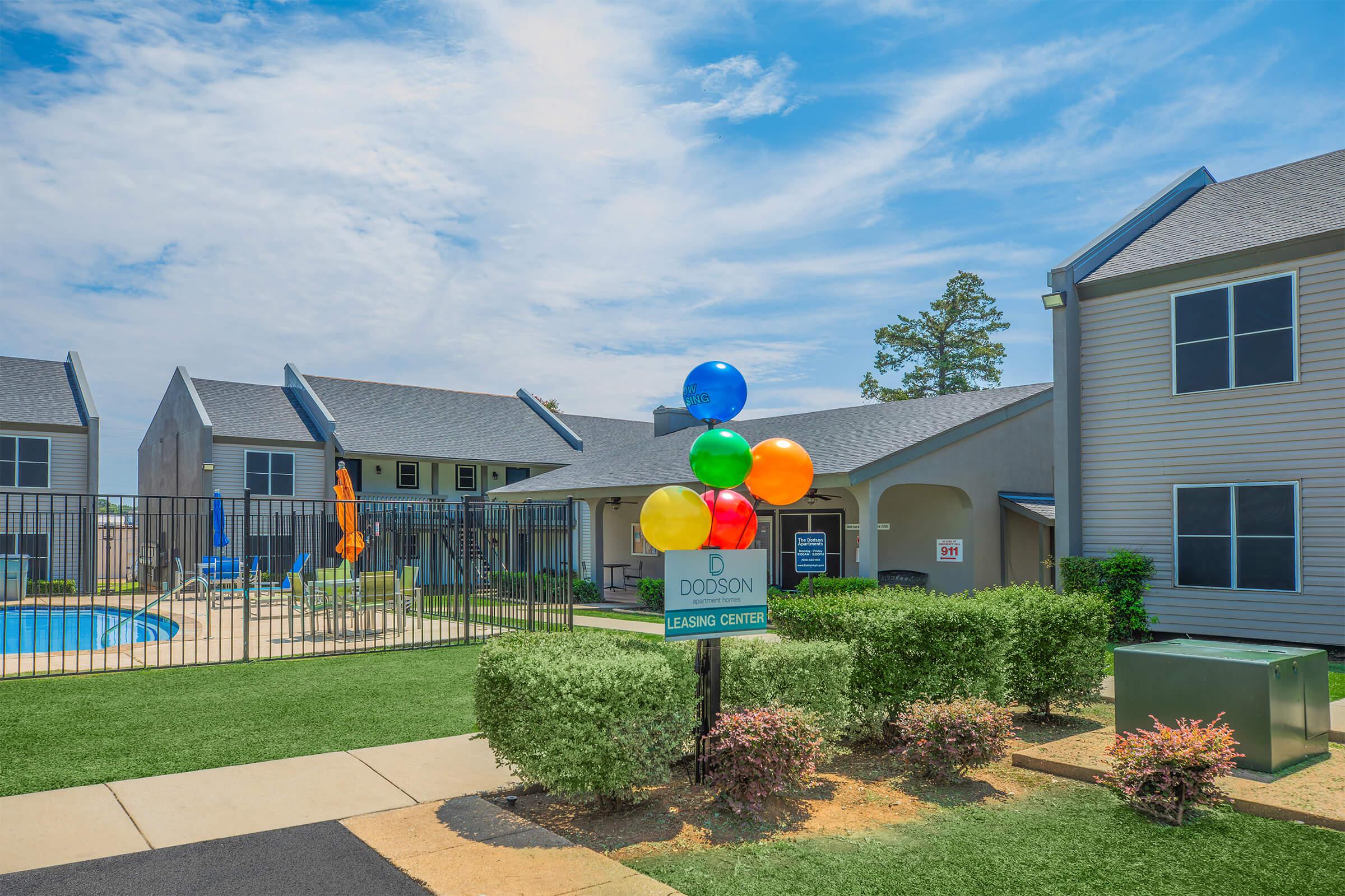 Leasing center sign at an apartment complex with colorful balloons nearby. In the background, a swimming pool surrounded by lounge chairs and umbrellas, along with several multi-story buildings and green grass under a bright blue sky.
