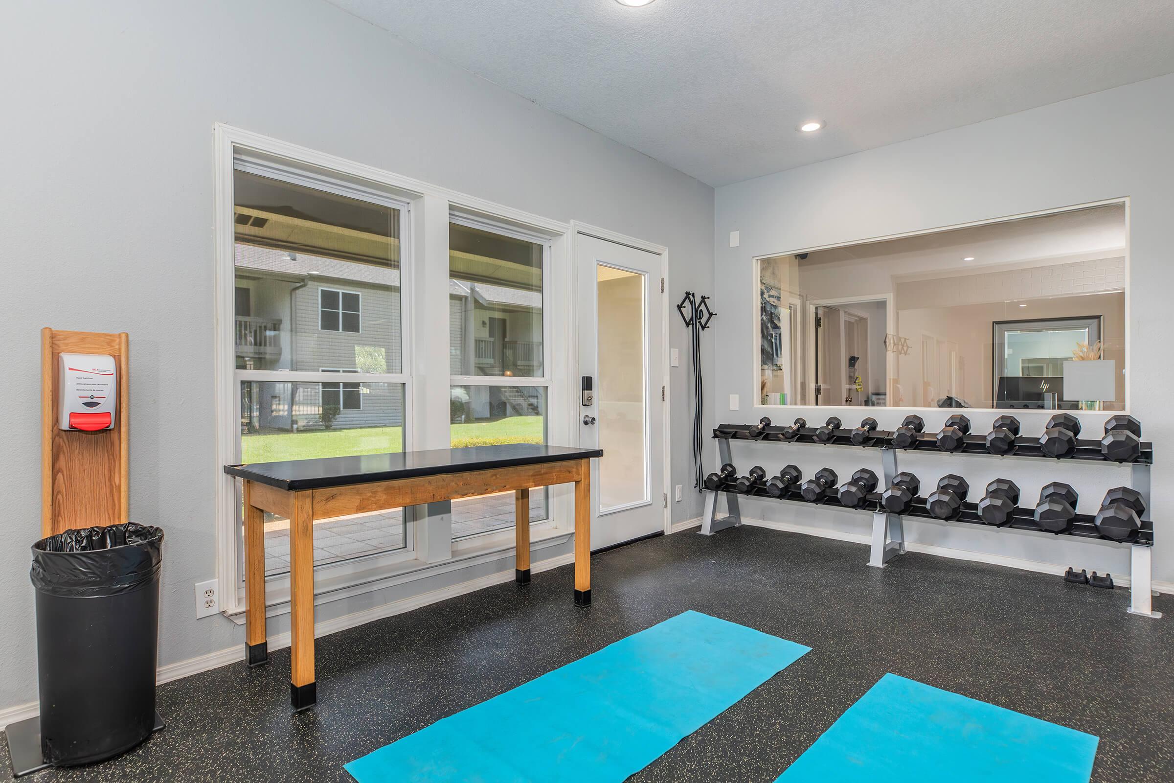 A modern gym interior featuring two blue exercise mats on a black rubber floor, a wooden table, a trash can, and a rack of dumbbells against the wall. Large windows provide natural light, and a mirror reflects the space, creating an open and welcoming workout environment.