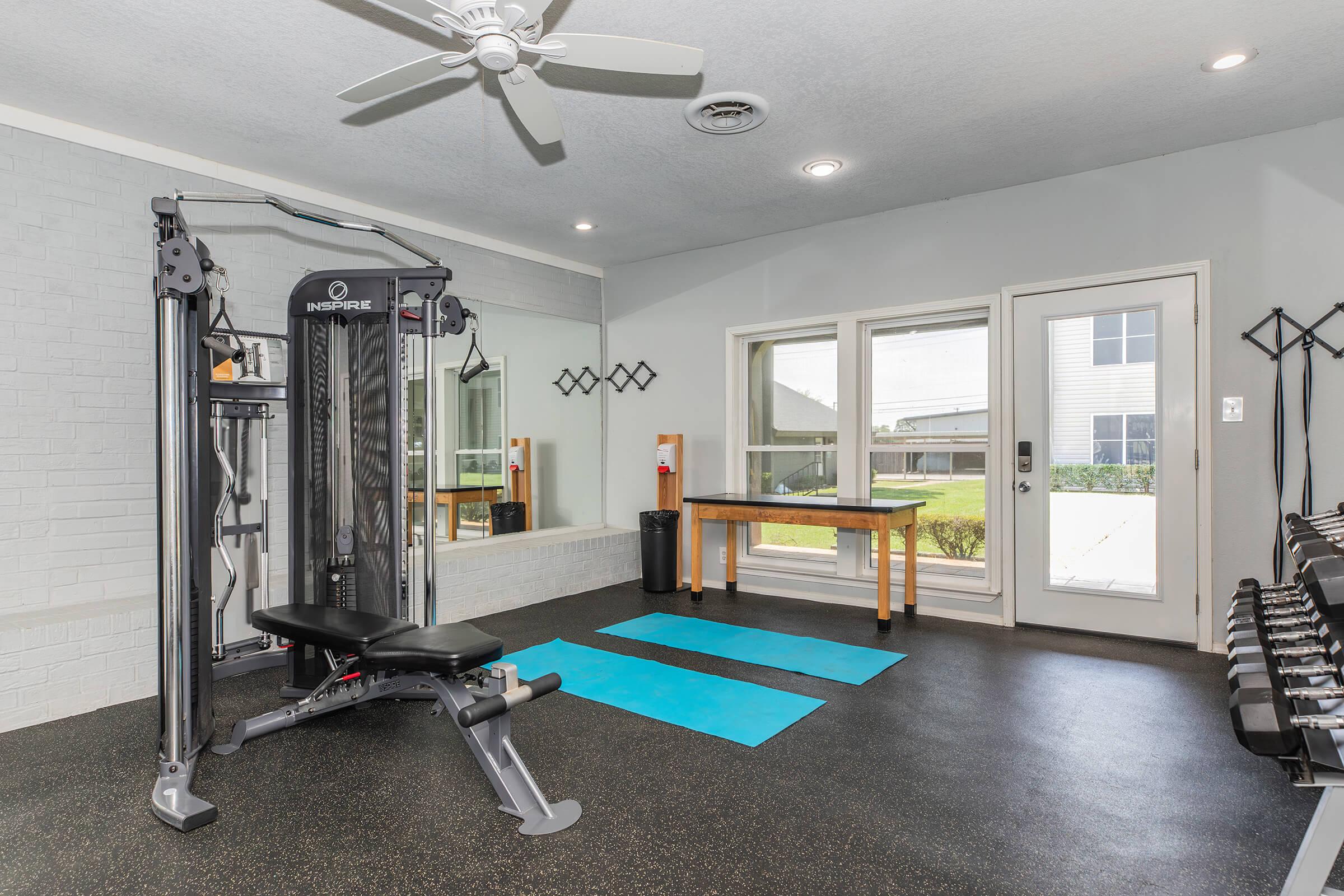 A modern home gym featuring a cable machine, two blue exercise mats on the floor, and a set of dumbbells. Large windows provide natural light, and there is a wood table near the entrance. The walls are light-colored, and a ceiling fan is installed.