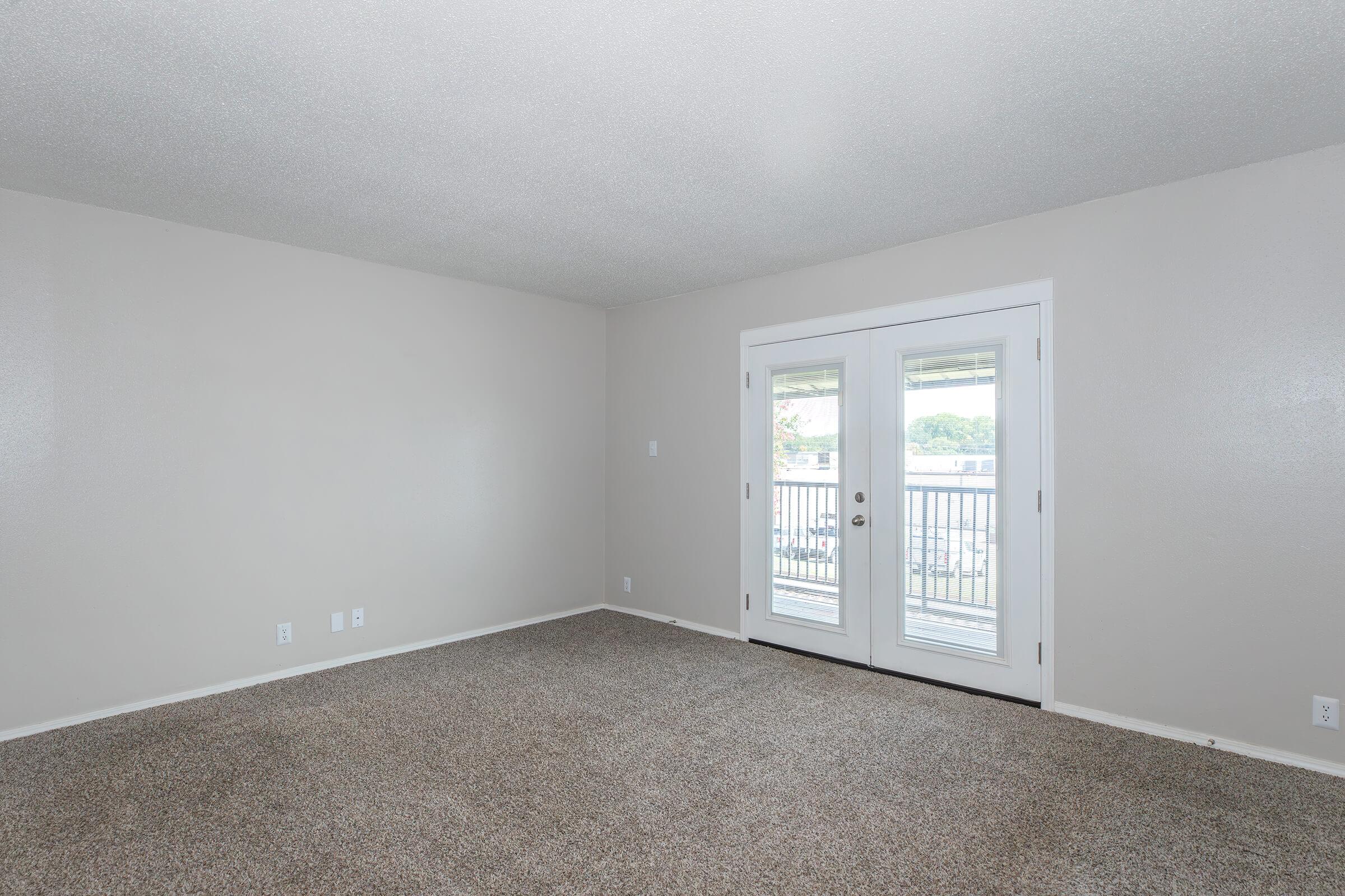 Empty room with light gray walls and carpeted floor. There are large sliding glass doors on one wall that lead to a small balcony, allowing natural light to enter. The room is unfurnished and spacious, creating a blank canvas for decoration.