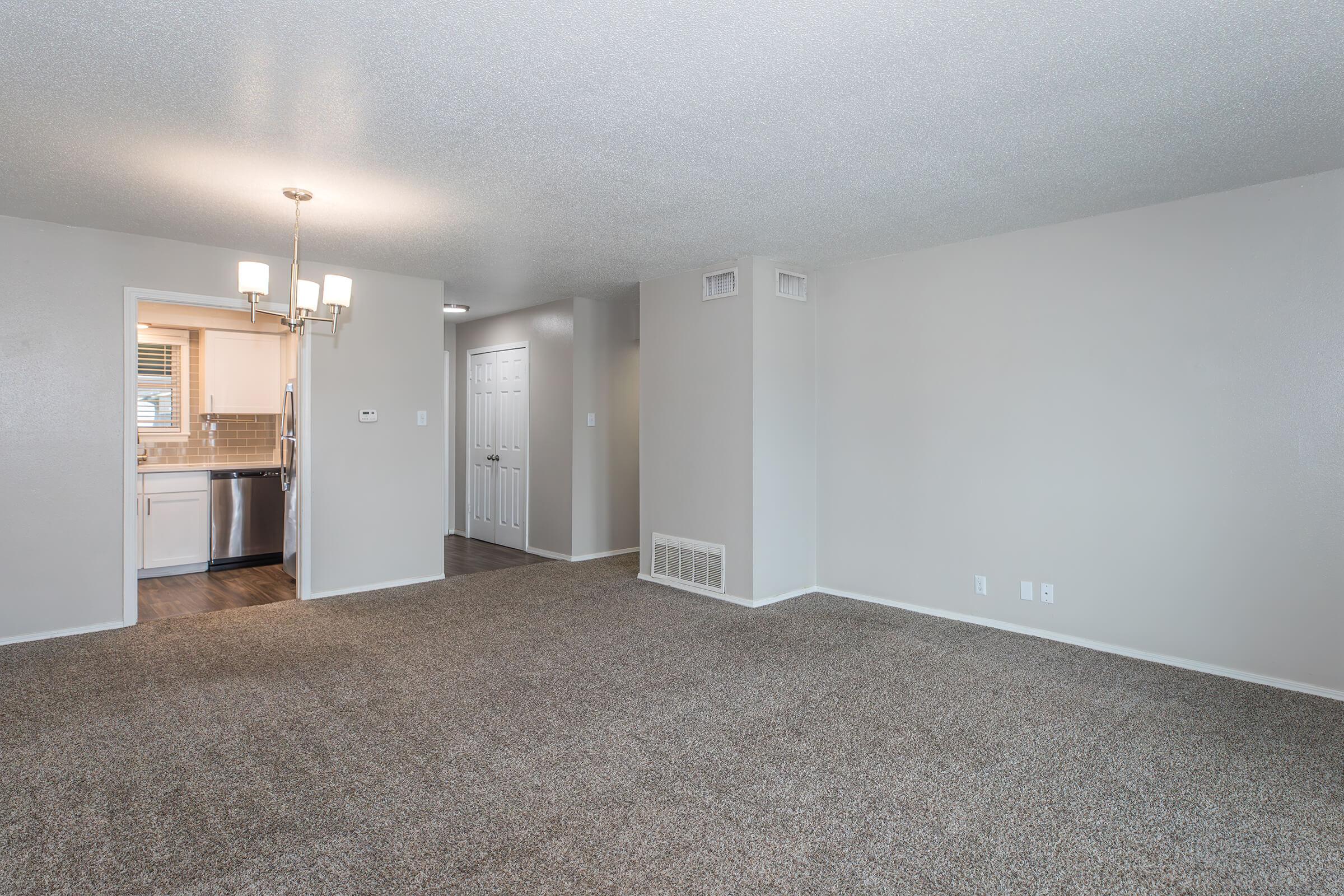 A spacious, empty living room with light gray walls and carpet. A doorway leads to a kitchen on the left, and there is a hallway with a closed door on the right. Overhead, a modern chandelier provides soft lighting, enhancing the welcoming atmosphere of the area.