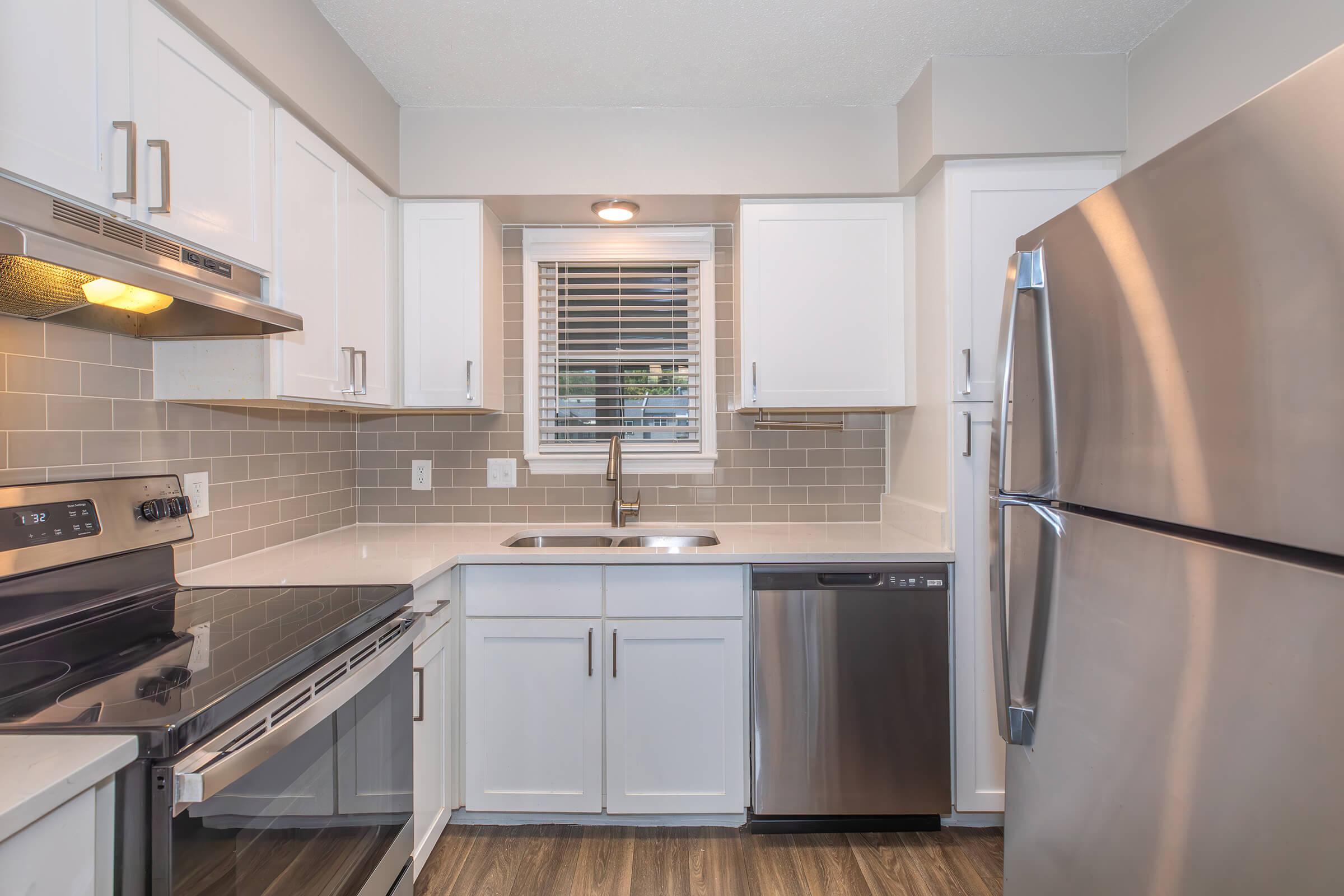 Modern kitchen featuring stainless steel appliances, including an oven, microwave, and refrigerator. The space has white cabinets, a light gray tile backsplash, a double sink under a window, and wood-style flooring, creating a bright and inviting atmosphere.