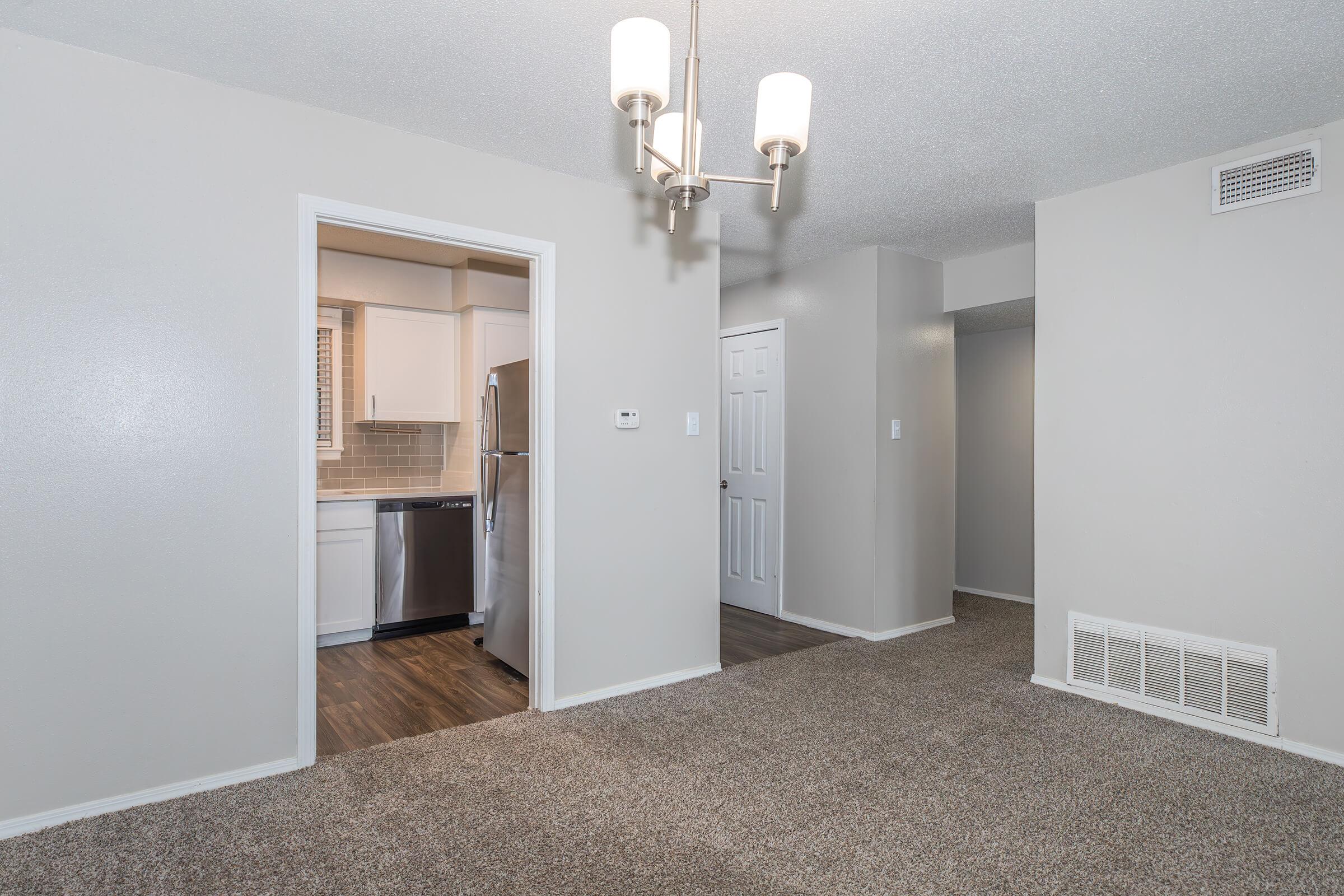 A well-lit interior view of an apartment, featuring a carpeted living area with a light fixture. To the left, a doorway leads to a modern kitchen with stainless steel appliances. Towards the right, there are two doors leading to separate rooms, with neutral-colored walls and flooring.