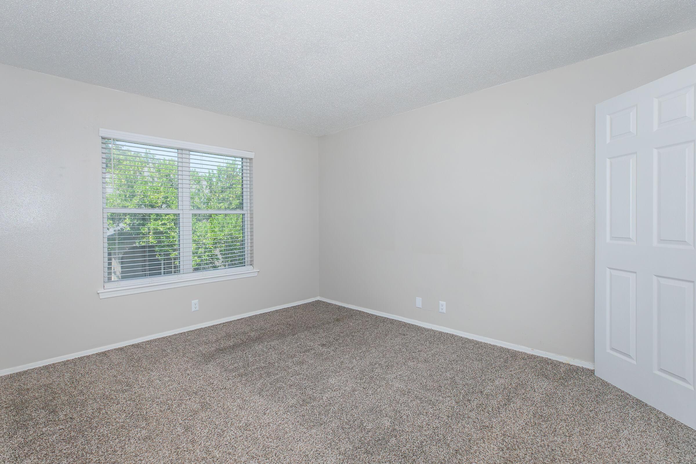 A clean, empty room with beige wall paint and light brown carpet. A window with white blinds allows natural light in, and there’s a white door on the right side. The space is uncluttered and ready for furnishings.