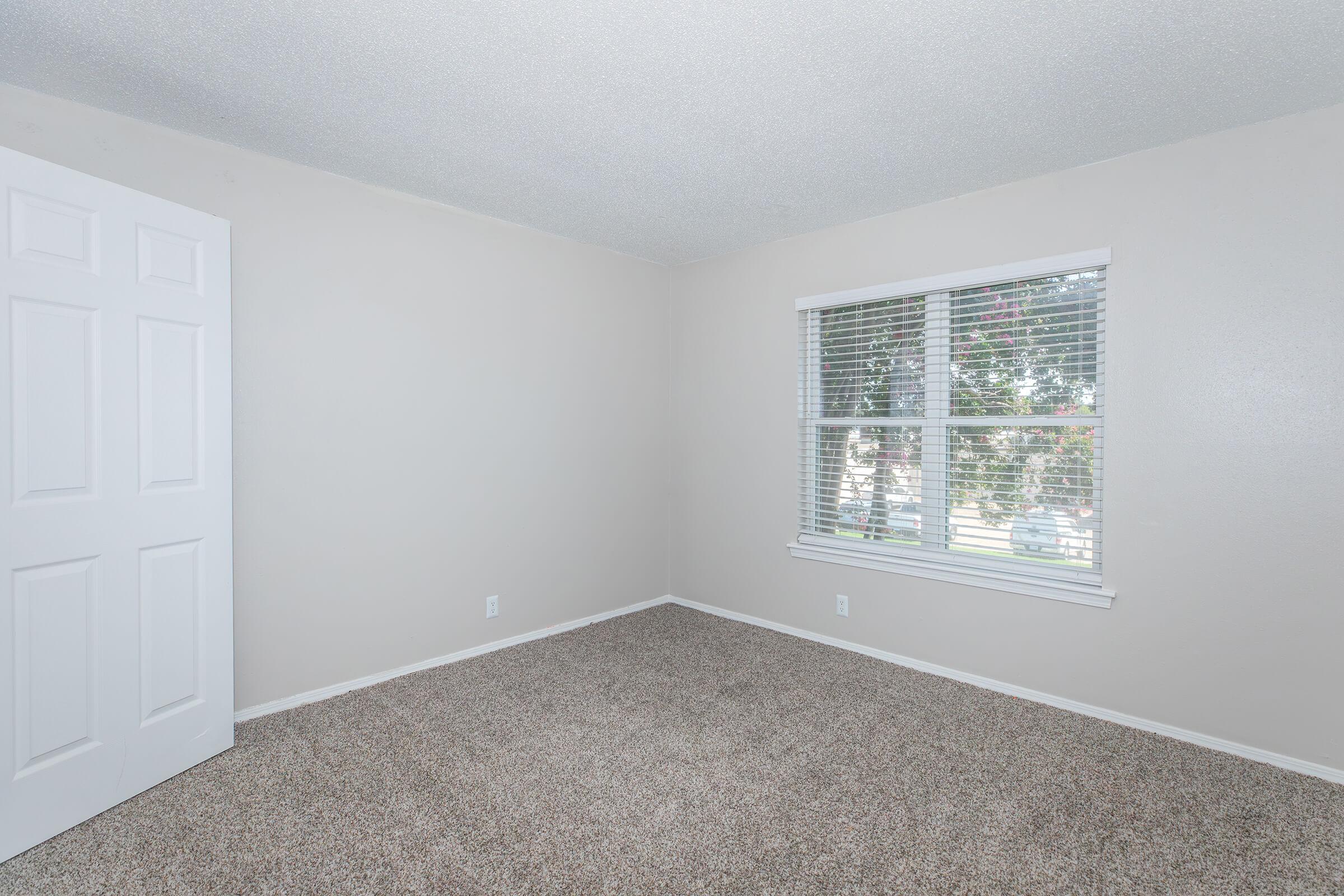 A vacant room with light beige walls and brown carpet. One corner features a white door leading to another area. A window with horizontal blinds allows natural light to enter, revealing a view of greenery outside. The space is unfurnished, providing a blank canvas for potential decorating.