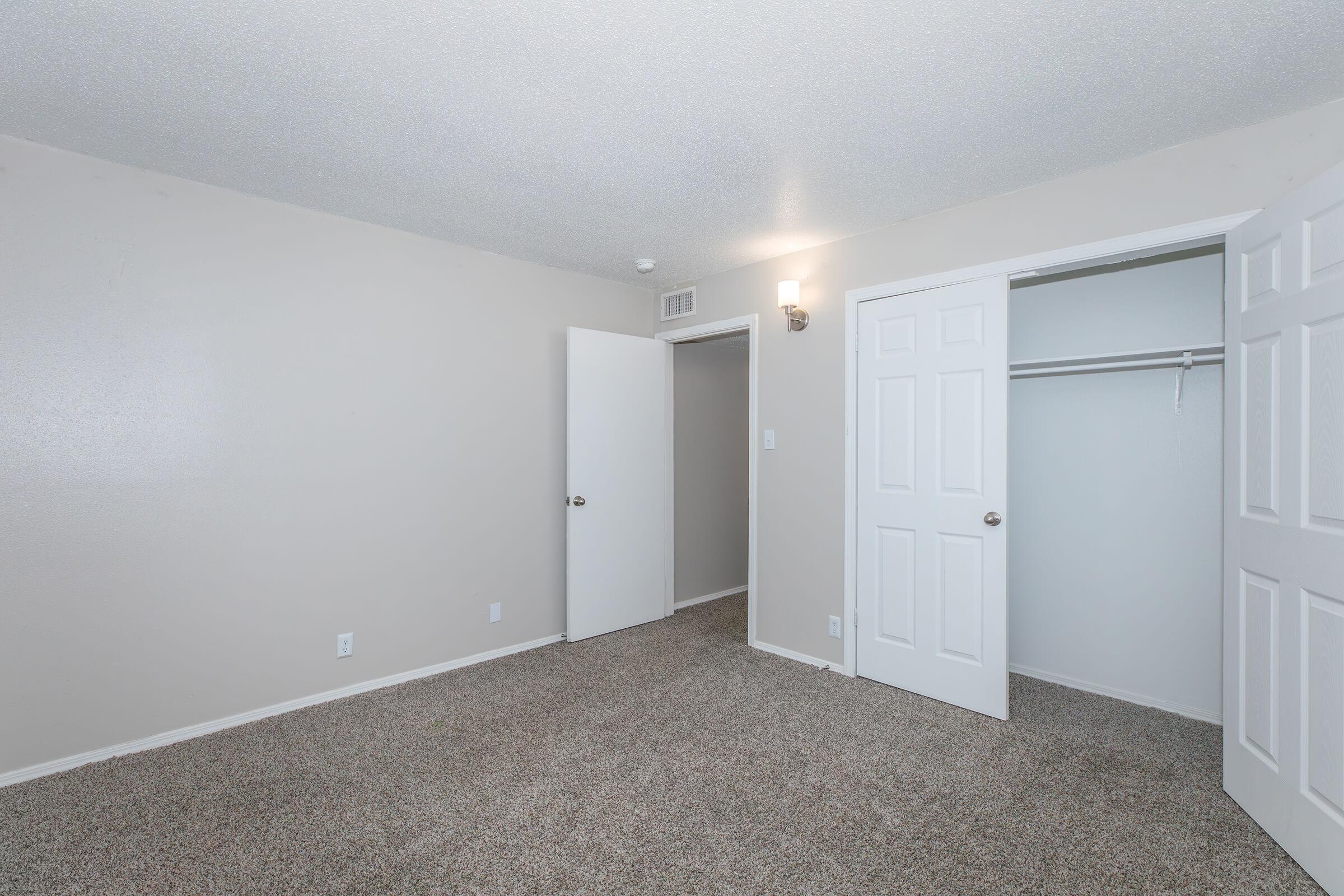 A vacant room with light beige walls and carpeted flooring. There are two white doors, one leading to a closet and the other potentially to another room. A wall sconce provides soft lighting. The overall atmosphere is empty and ready for furnishing.