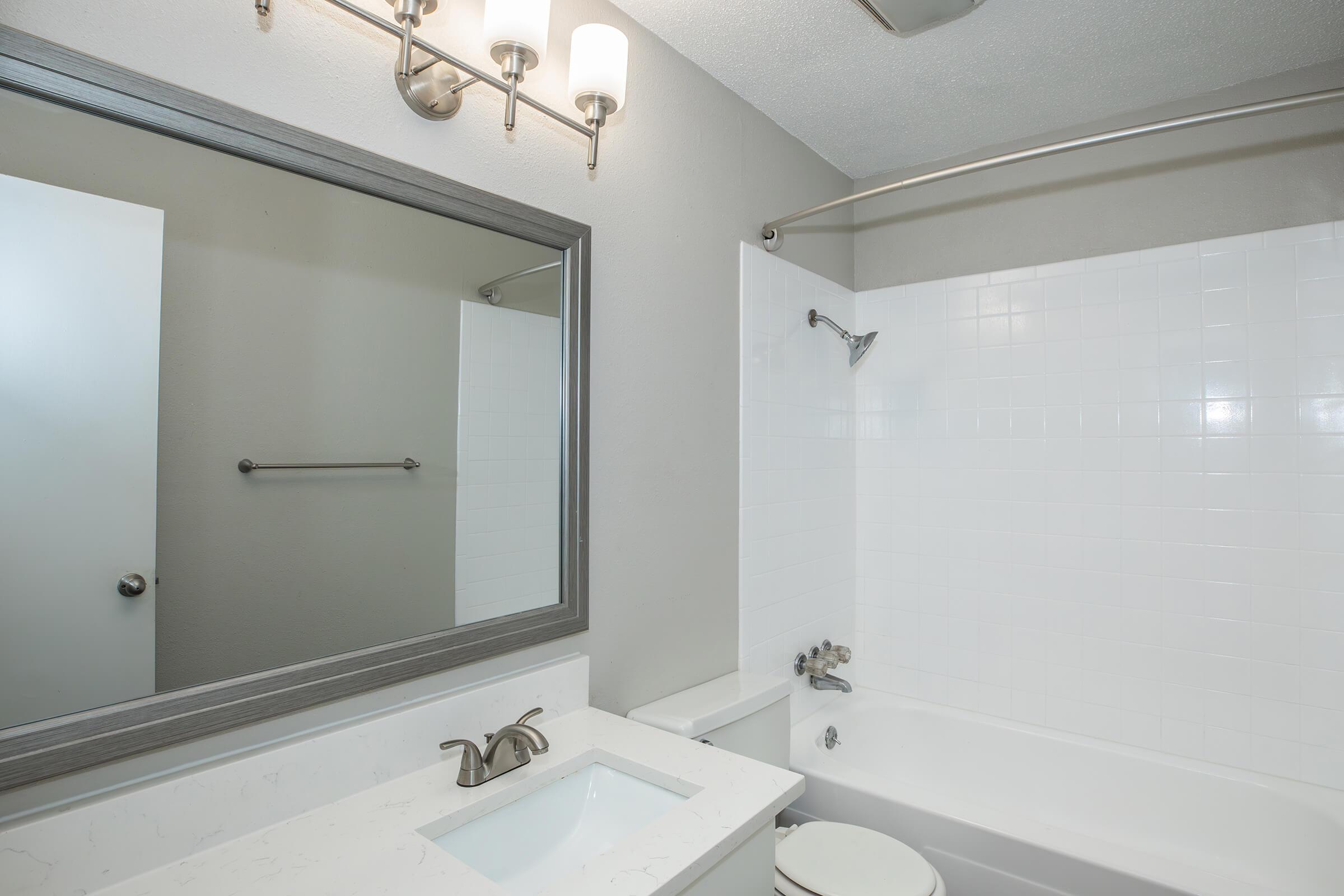 A modern bathroom featuring a white bathtub with a shower, a sleek sink with a faucet, and a large mirror above. The walls are painted light gray, and there is a towel rack mounted on the wall. Natural light illuminates the space, creating a clean and fresh ambiance.