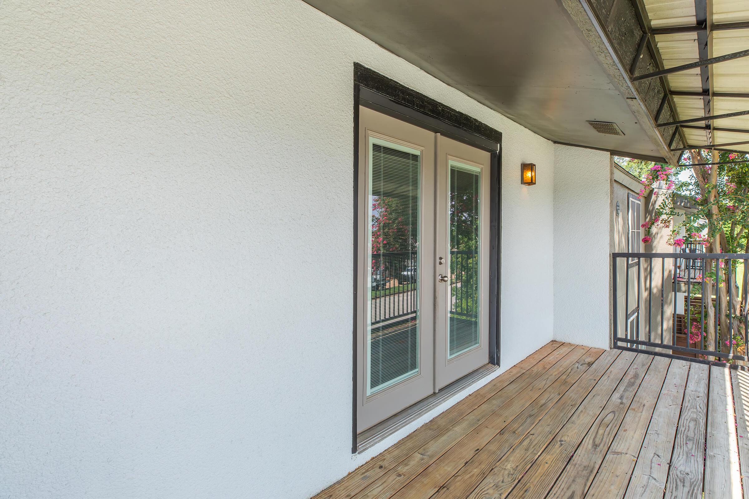 Exterior view of a balcony with a wooden floor, featuring a pair of double doors with glass panels leading to an interior space. The wall is painted white, and there is a small wall-mounted light fixture beside the doors. A railing can be seen, along with greenery in the background.