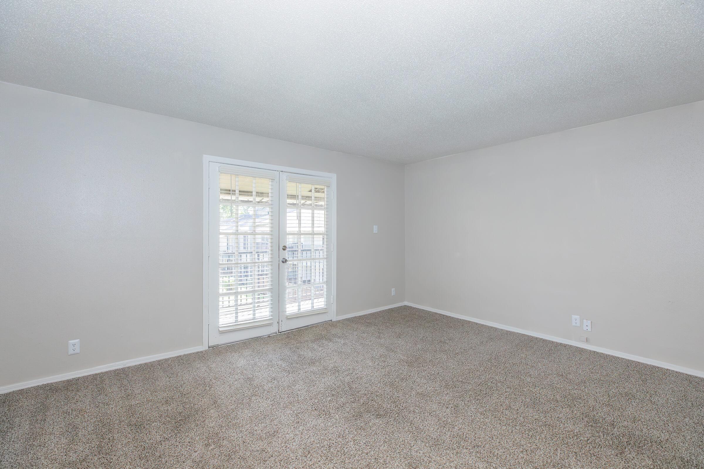 A spacious, empty living room with light beige carpet and neutral-colored walls. Large sliding glass doors with white shutters lead to a balcony, allowing natural light to brighten the area. The room has no furniture, creating a clean and open atmosphere.