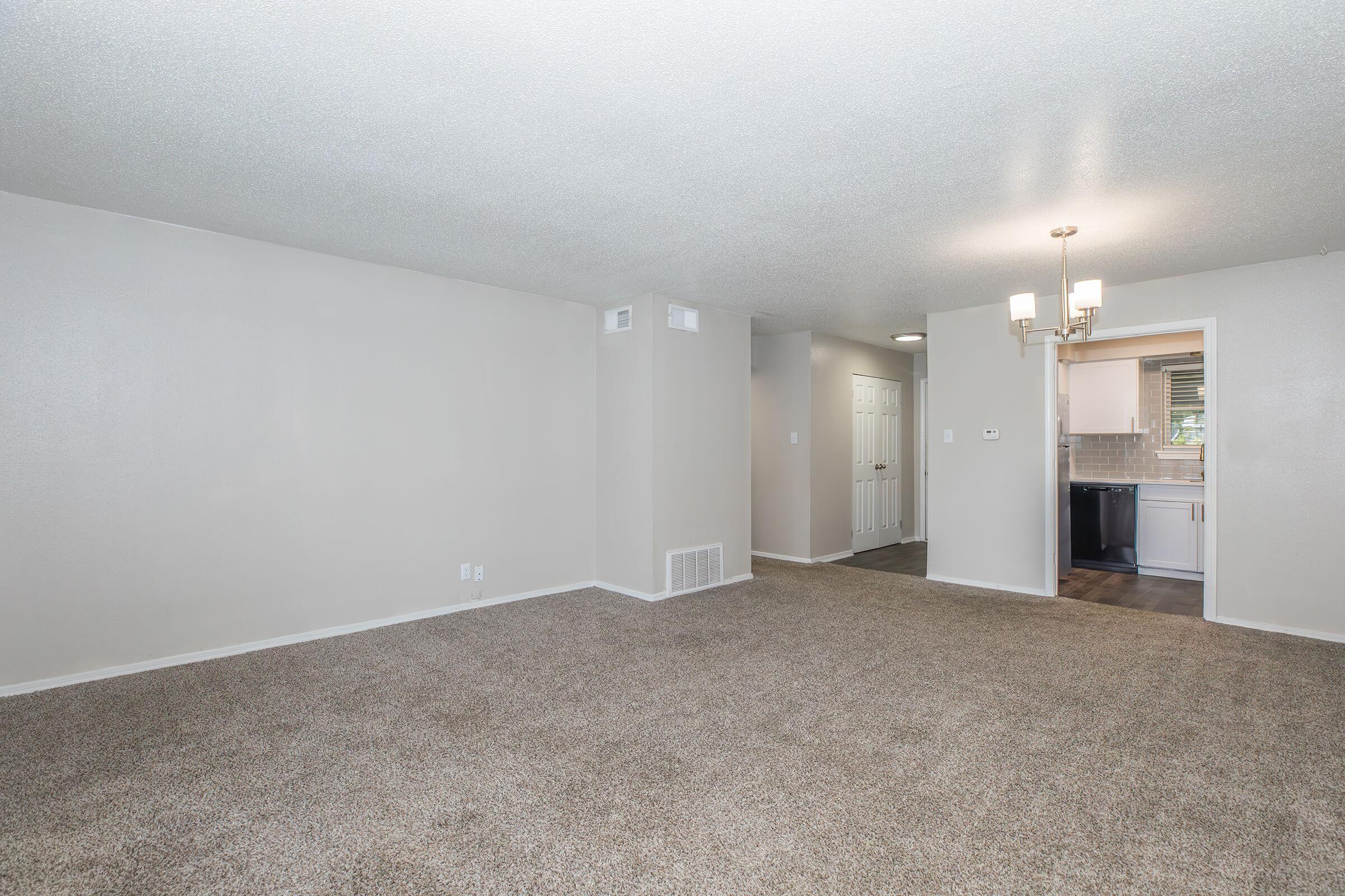 Spacious living room with light-colored walls and plush carpet. The area features a doorway leading to a kitchen and another to a hallway. A modern chandelier hangs from the ceiling, adding a touch of elegance to the neutral-toned space, which is well-lit by natural light.