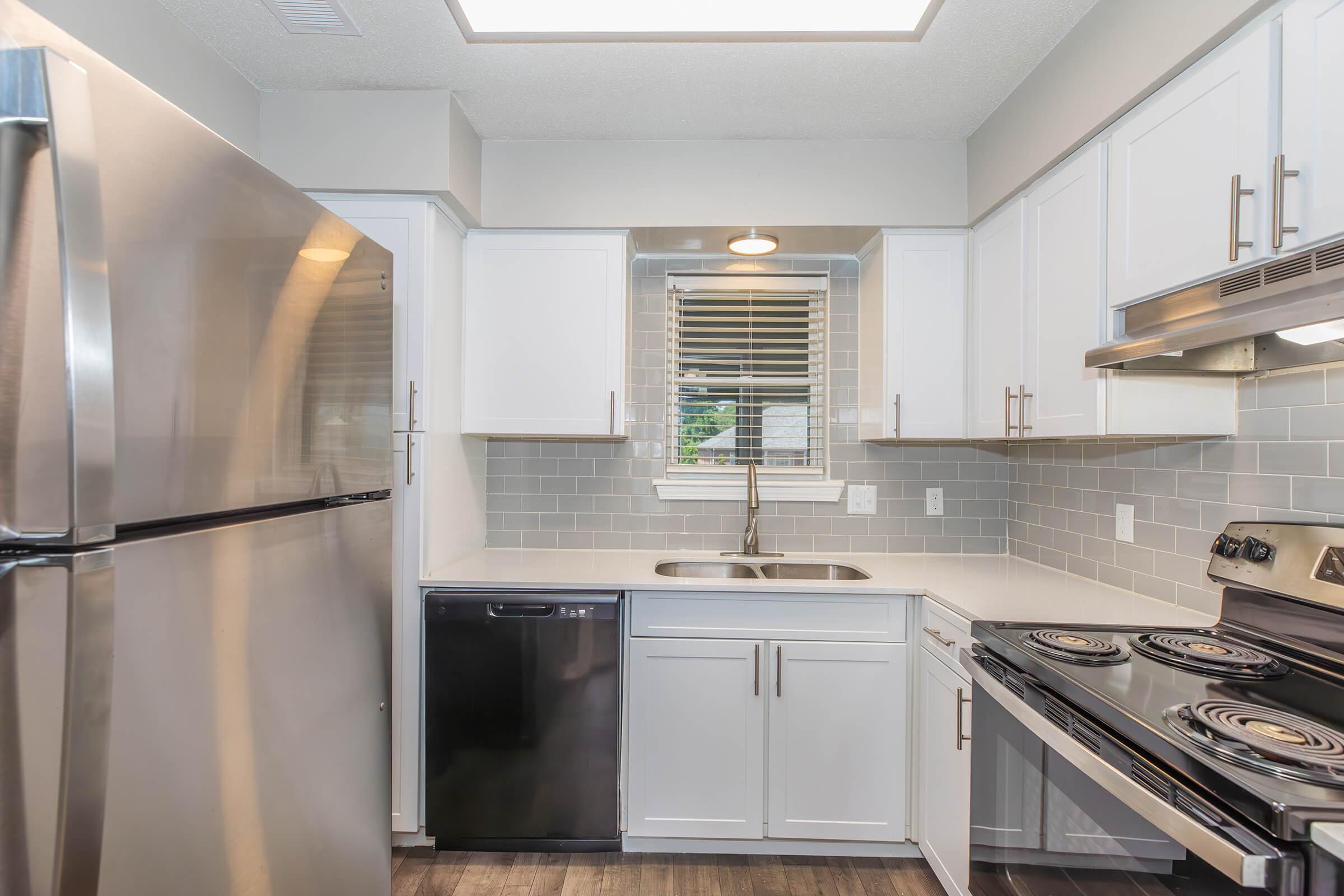 A modern kitchen with stainless steel appliances, including a refrigerator, dishwasher, and stove. The cabinetry is white, complemented by a light grey tiled backsplash. Natural light comes in through a window above the sink, and the countertop is a clean, light color, enhancing the spacious feel of the kitchen.
