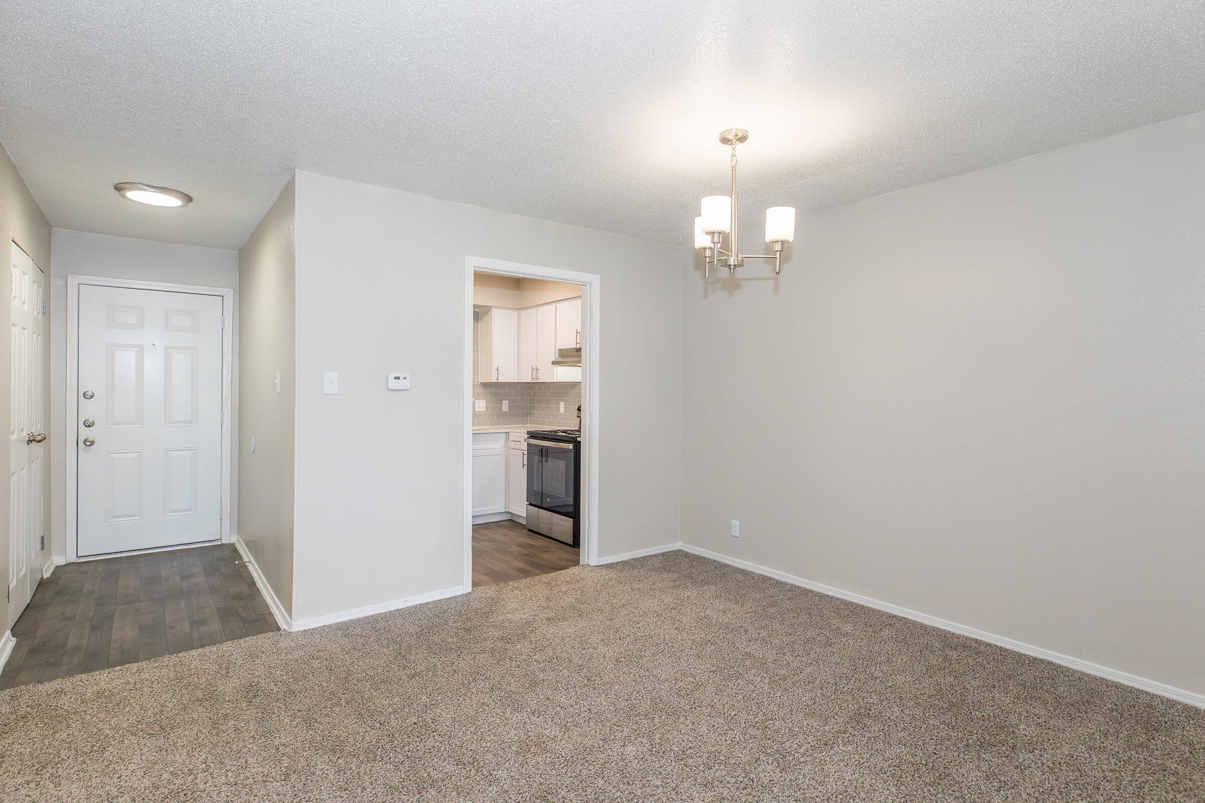 A well-lit living area featuring beige walls and plush carpet. On the left, a white door leads to an entrance. In the background, a doorway opens to a kitchen with modern cabinetry and appliances. Ceiling lights and a chandelier add to the ambiance, creating a cozy and inviting atmosphere.