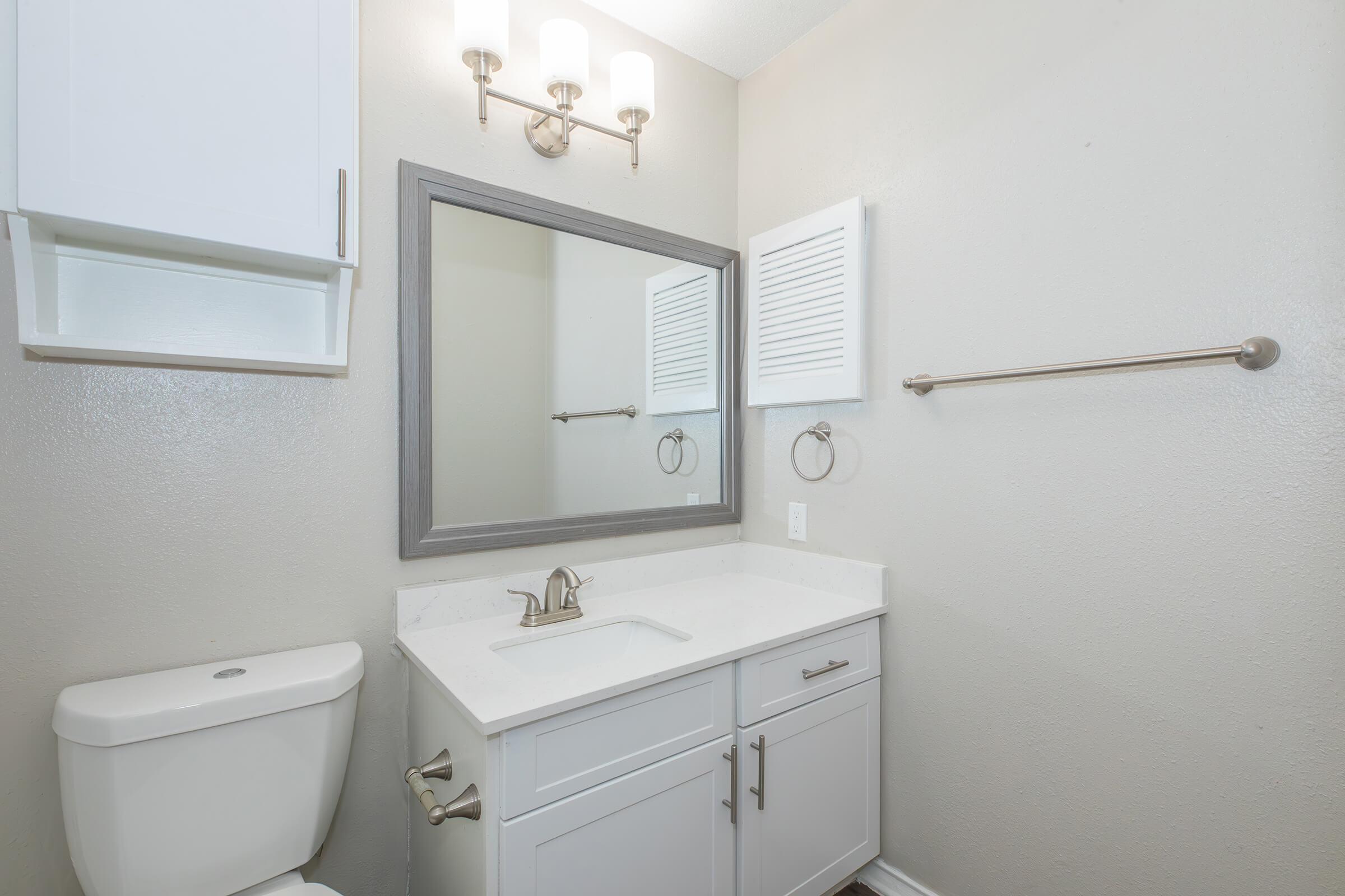 A modern bathroom featuring a white vanity with a sink, a rectangular mirror framed in gray, and stylish lighting fixtures above. The walls are painted in a light color, and there is a toilet and towel rack visible. The overall design is clean and contemporary.