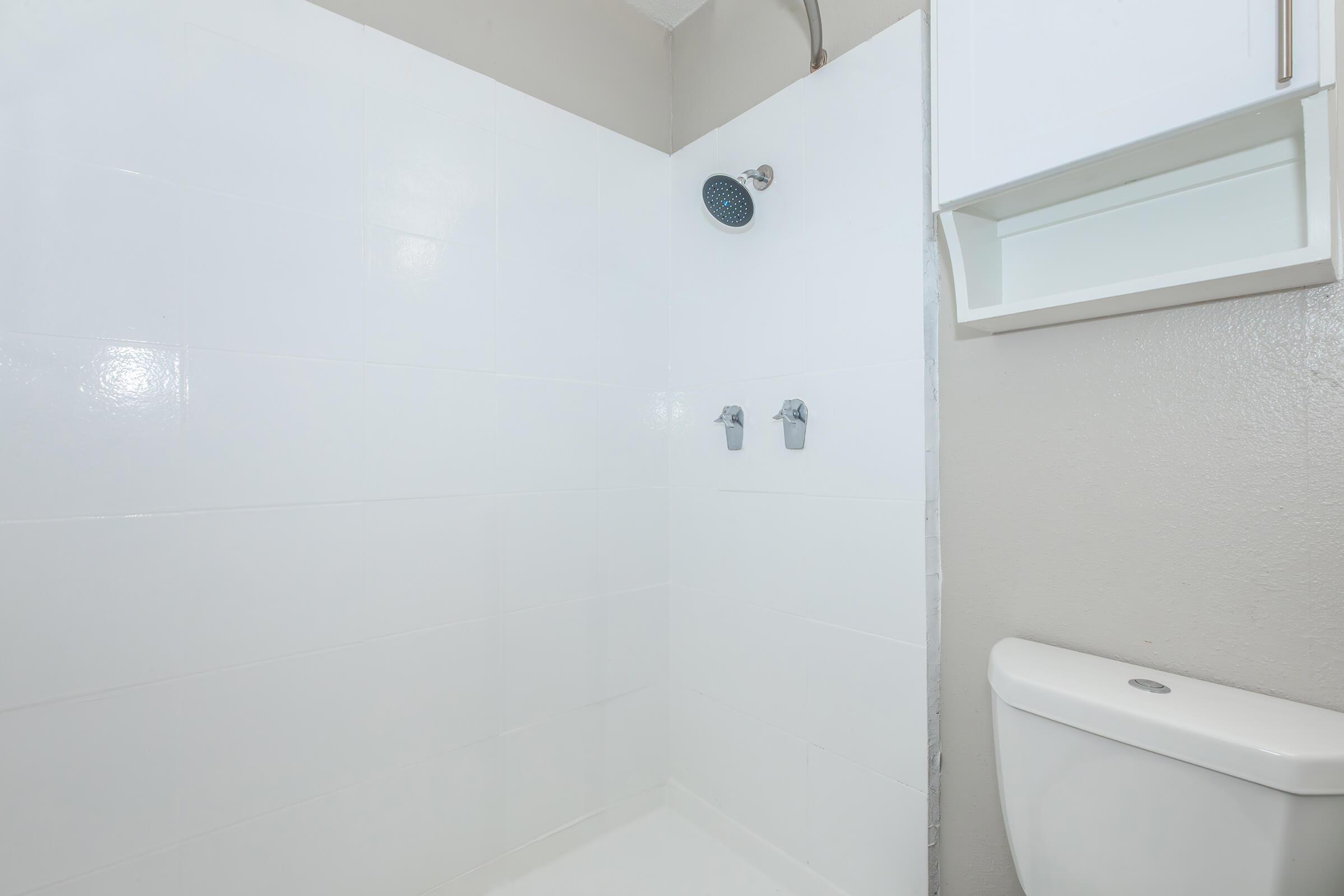 A clean and modern bathroom shower area featuring white tiled walls, a rain showerhead on the left, and two silver faucet handles. A white toilet is situated on the right side, with a small shelf above the shower area for storage. The overall aesthetic is minimal and bright.