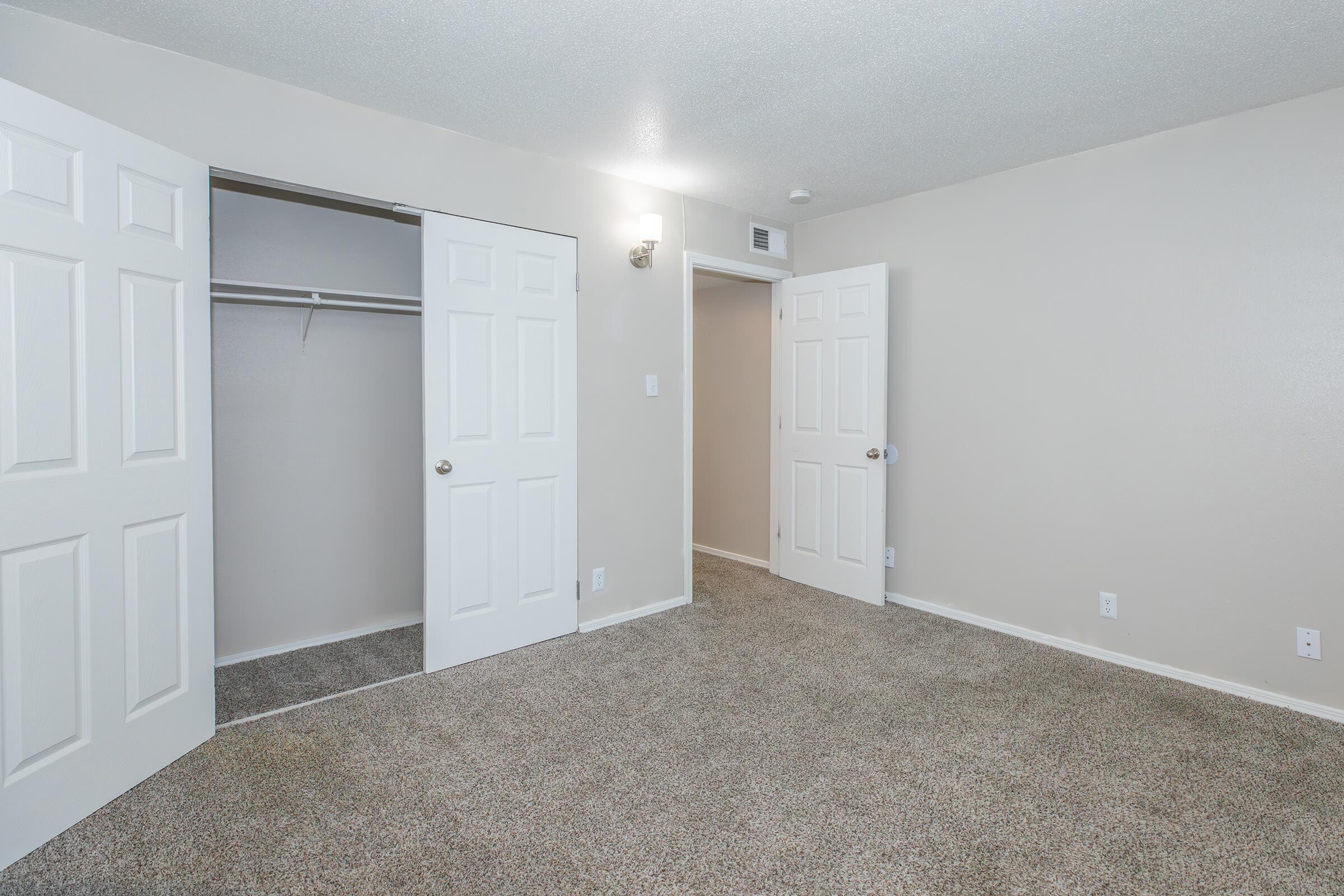 A well-lit, empty room with light beige walls and carpet. Two closed doors lead to closets with a single hanging rod. The room features a wall-mounted light fixture and a clear view into another doorway, suggesting additional space. The overall atmosphere is clean and spacious.