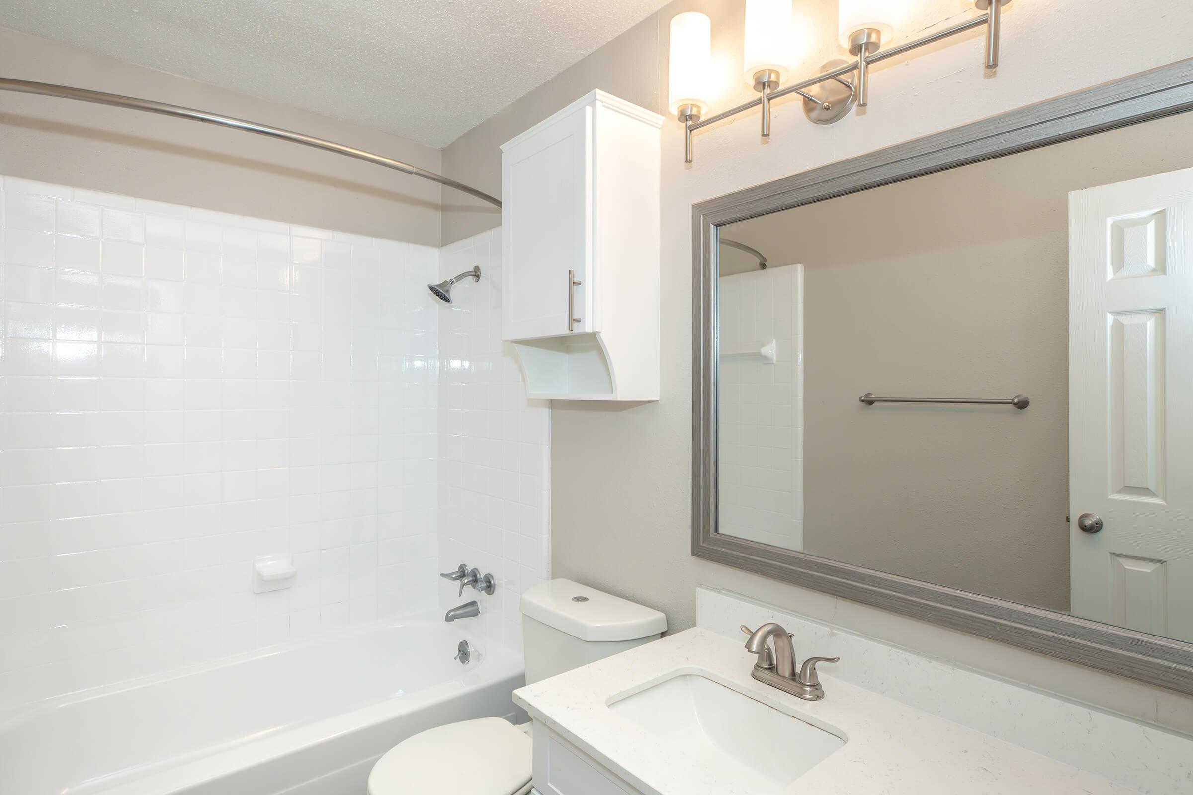 A clean, modern bathroom featuring a white bathtub with a showerhead, a light-colored countertop with a sink, a large mirror, and a light fixture above. The walls are painted in a neutral tone, and there is a towel bar next to a closed door.
