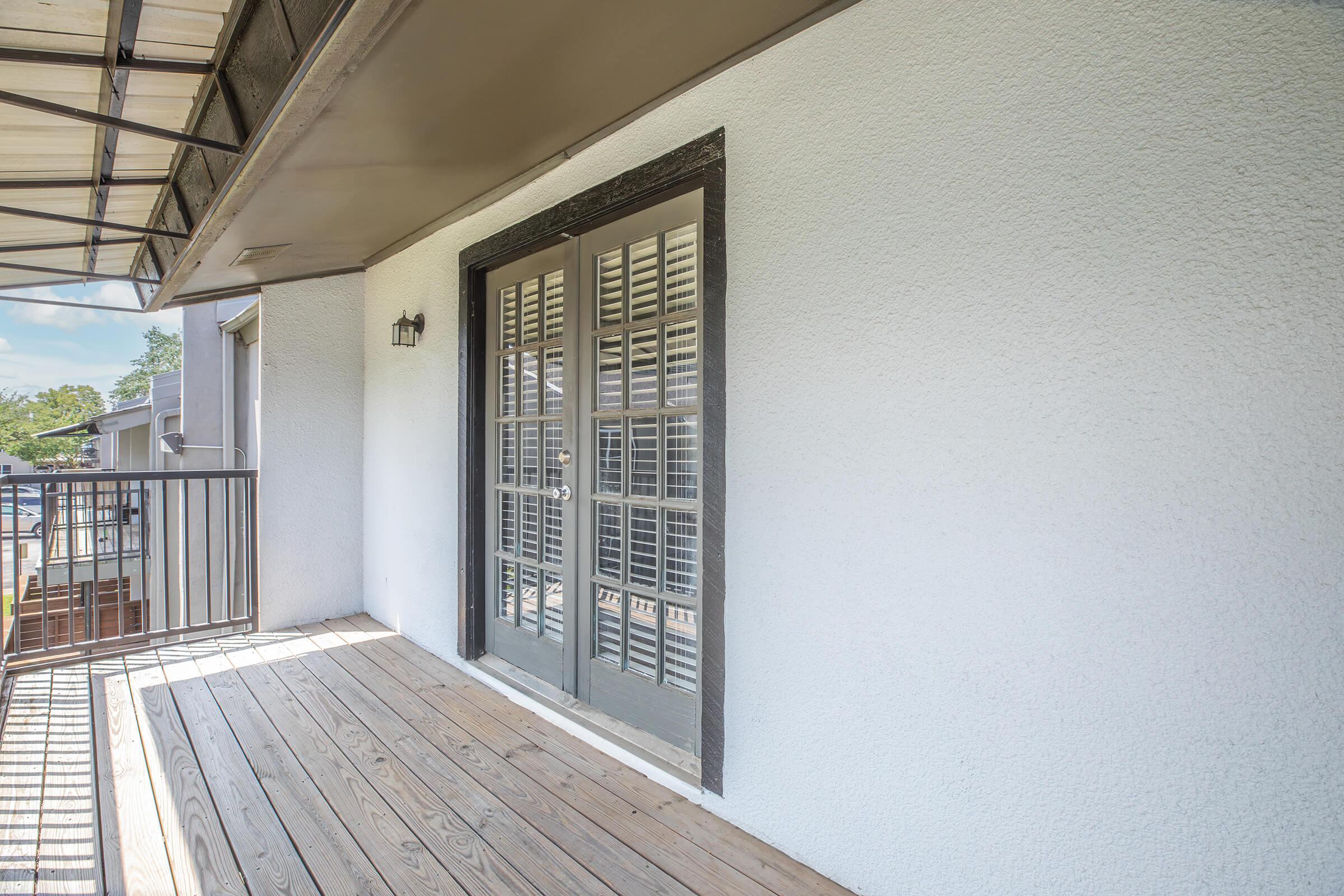 A balcony with a wooden floor, featuring a pair of closed French doors with shutters. The walls are painted white, and there is a lamp next to the door. The balcony is under a covered awning with a clear blue sky visible in the background.