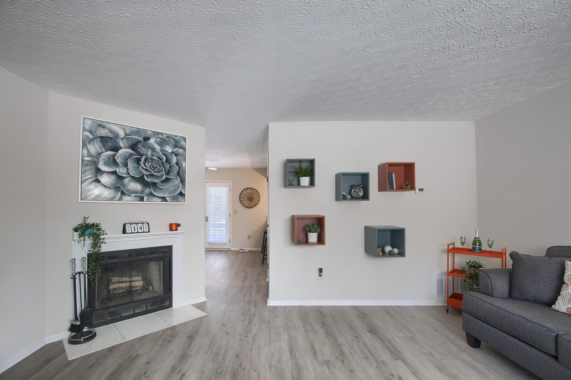A modern living room featuring a large succulent wall art above a fireplace. The room has light-colored walls and a wooden floor, with decorative shelves in the corner displaying plants and knickknacks. A gray sofa is positioned nearby, creating a cozy atmosphere. Natural light enters from a nearby window.