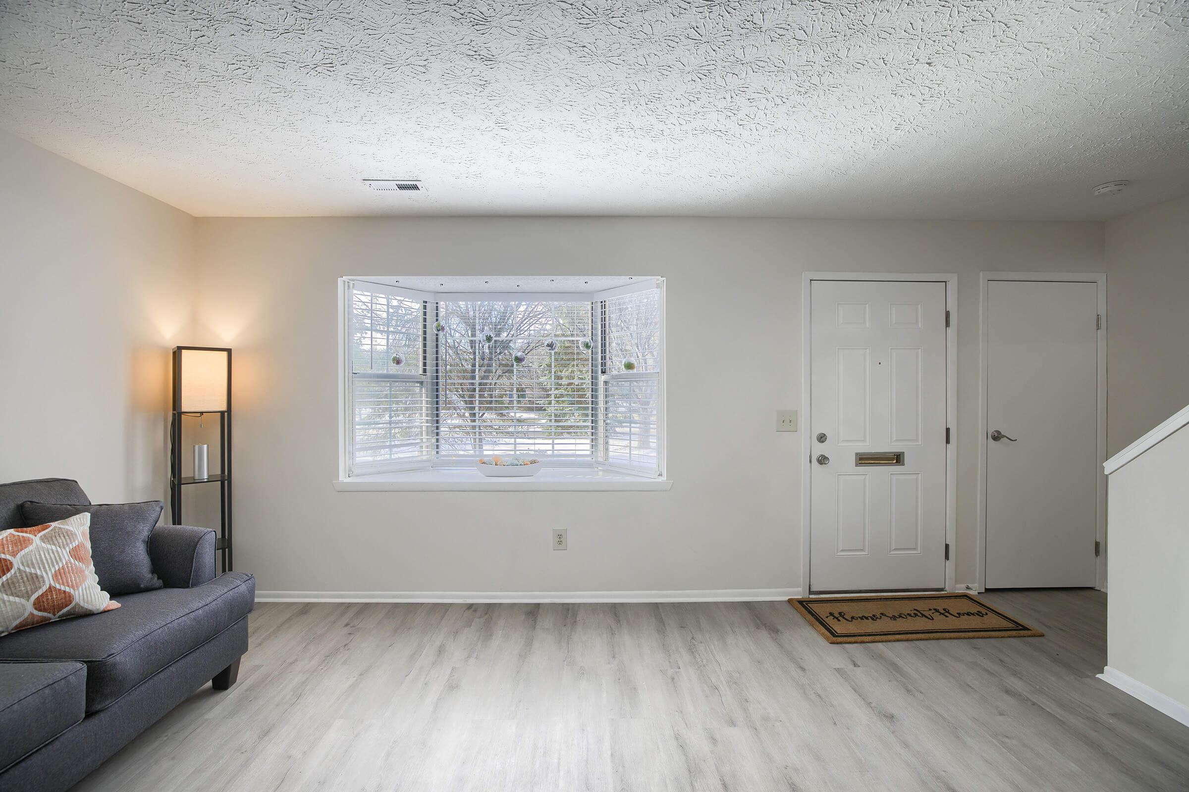 A clean and bright living space featuring a gray couch with decorative pillows, a bay window letting in natural light, a lamp beside the couch, and a door leading to another room. The floor is light-colored laminate, and there’s a welcome mat near the entrance.