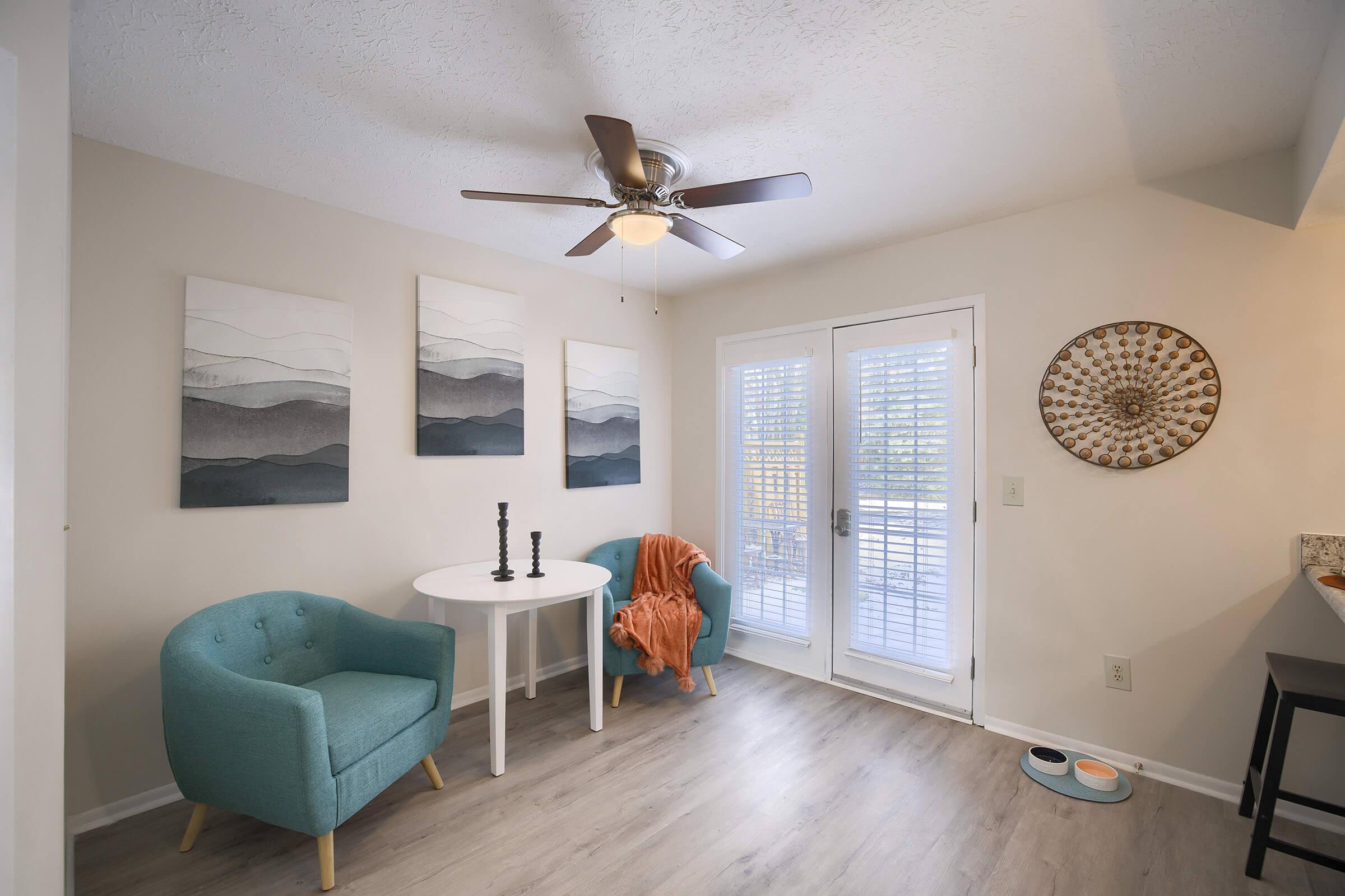 A cozy dining nook featuring a round white table with two turquoise chairs, a decorative orange throw draped over one chair, and three framed art pieces on the wall. Sunlight streams through glass doors leading to an outdoor space, and a decorative wall plate is visible nearby. A modern ceiling fan completes the space.