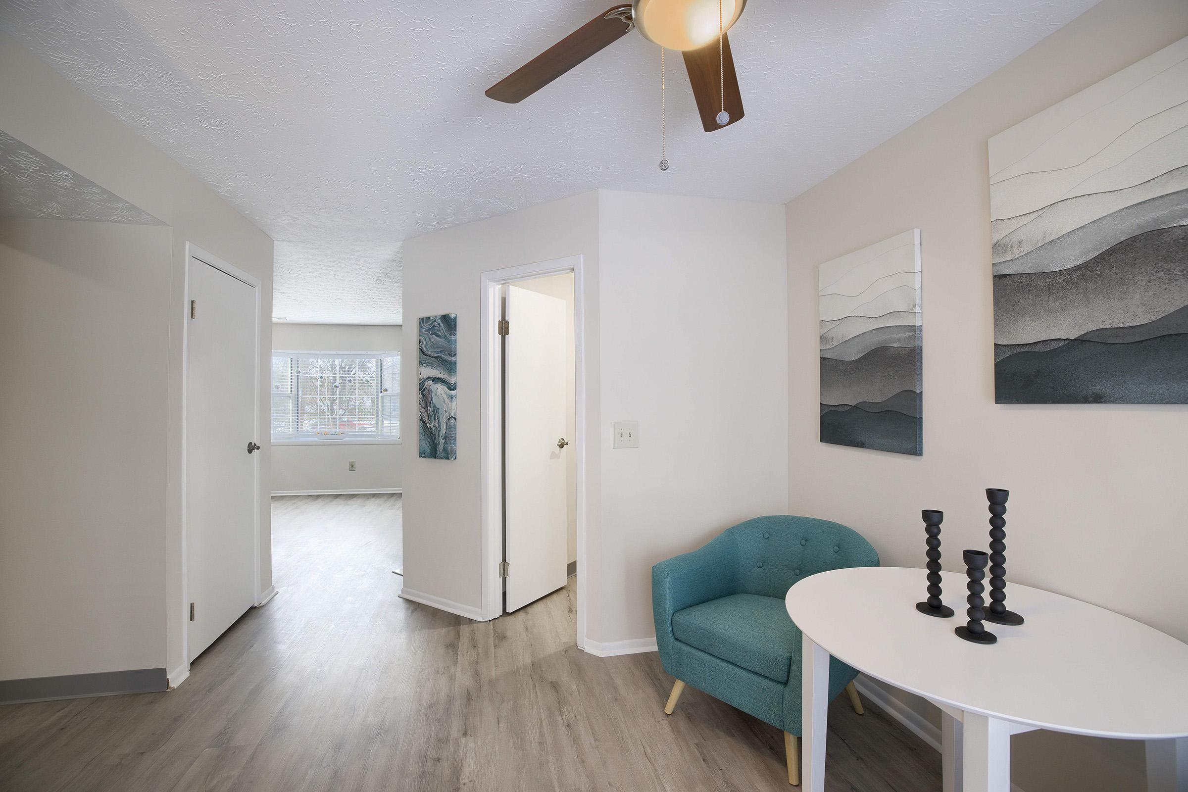 A well-lit interior space featuring a small table with black candlesticks and a teal chair. The walls are painted in neutral tones, with two abstract art pieces hanging above the table. A doorway leads to another room, and there is a ceiling fan visible. Large windows provide natural light.
