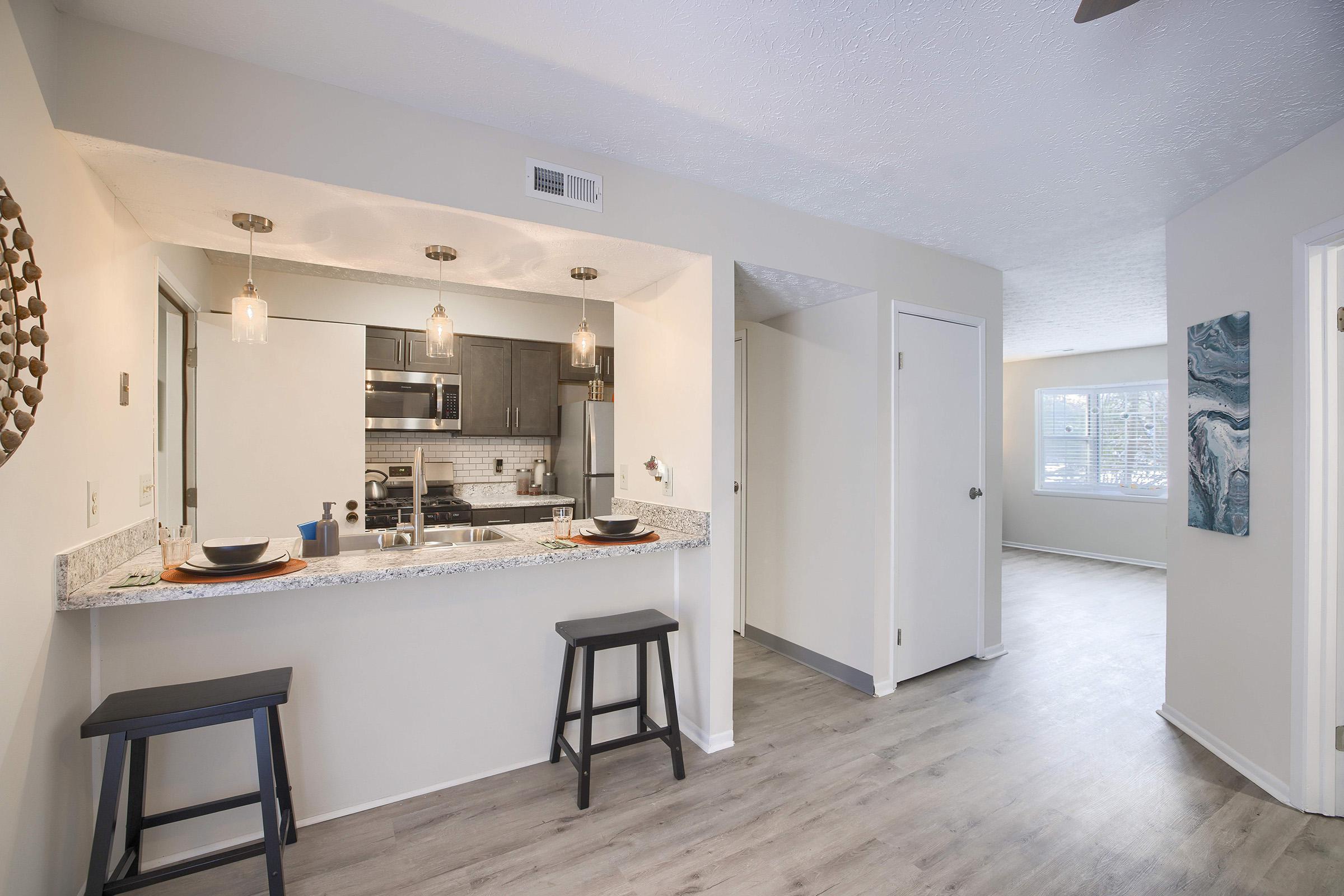 Bright, modern kitchen and dining area featuring a granite countertop with two stools. The kitchen has stainless steel appliances and pendant lighting. Soft, neutral walls and stylish decor enhance the open layout, leading to a sunlit living room with large windows. Fresh and inviting atmosphere throughout.