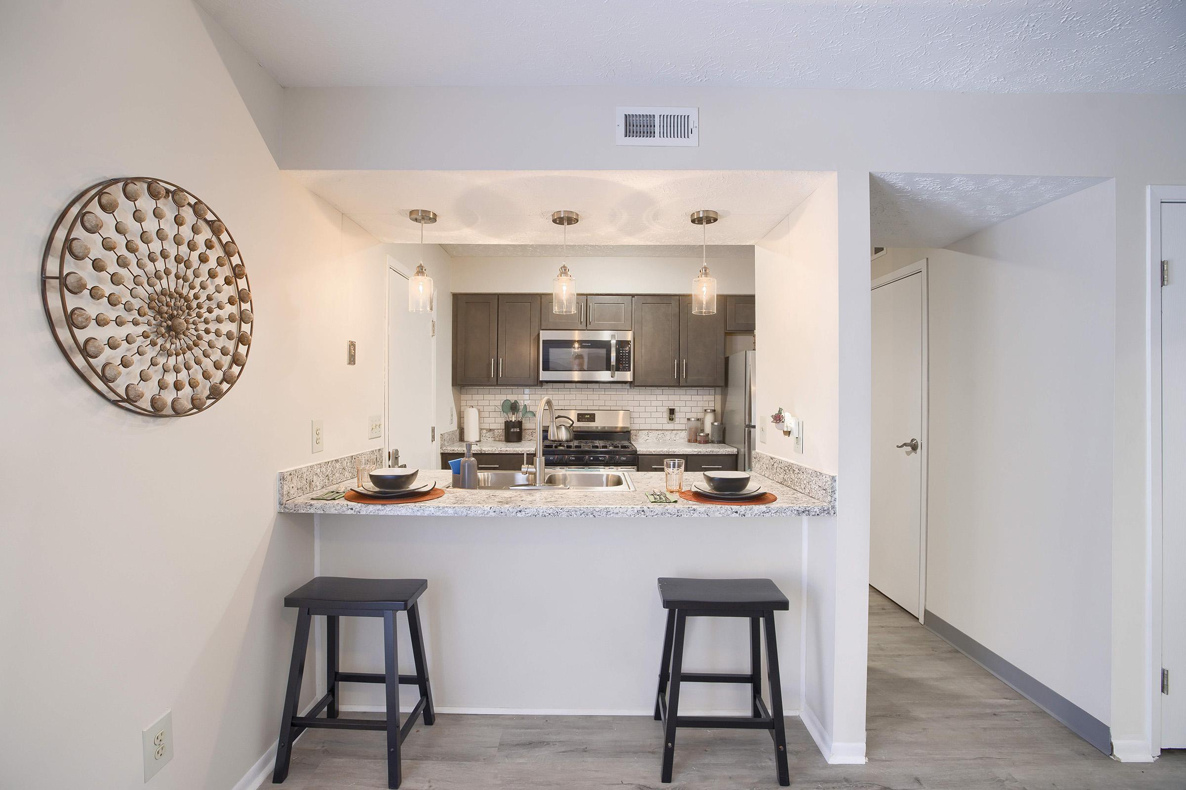 A modern kitchen with dark wood cabinets and stainless steel appliances. A granite countertop with two black stools is in the foreground, complemented by decorative elements on the wall. Soft lighting from pendant lamps enhances the inviting atmosphere. A doorway on the right leads to another room.