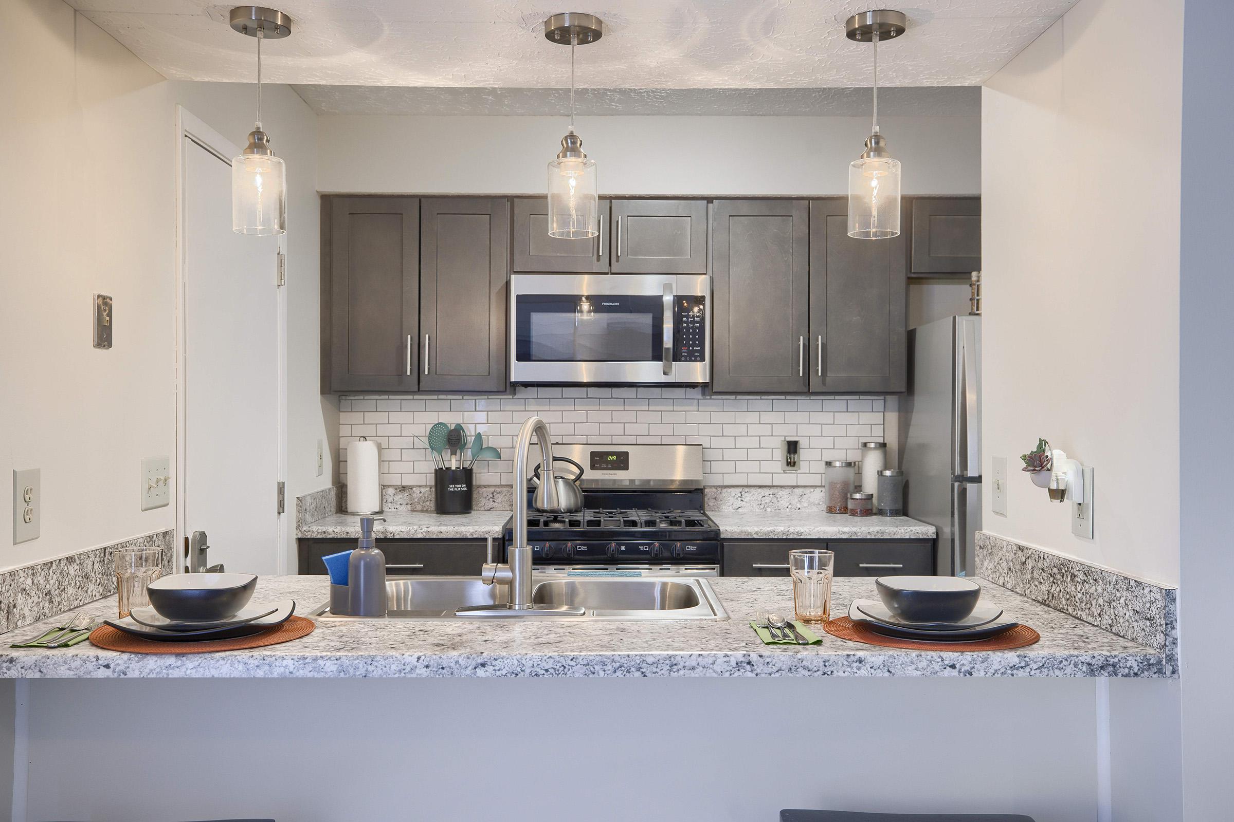 Modern kitchen featuring granite countertops, stainless steel appliances including a microwave and gas stove, and pendant lighting. The kitchen island is set with two place settings, including dishes and glasses, and there are decorative elements on the countertops.