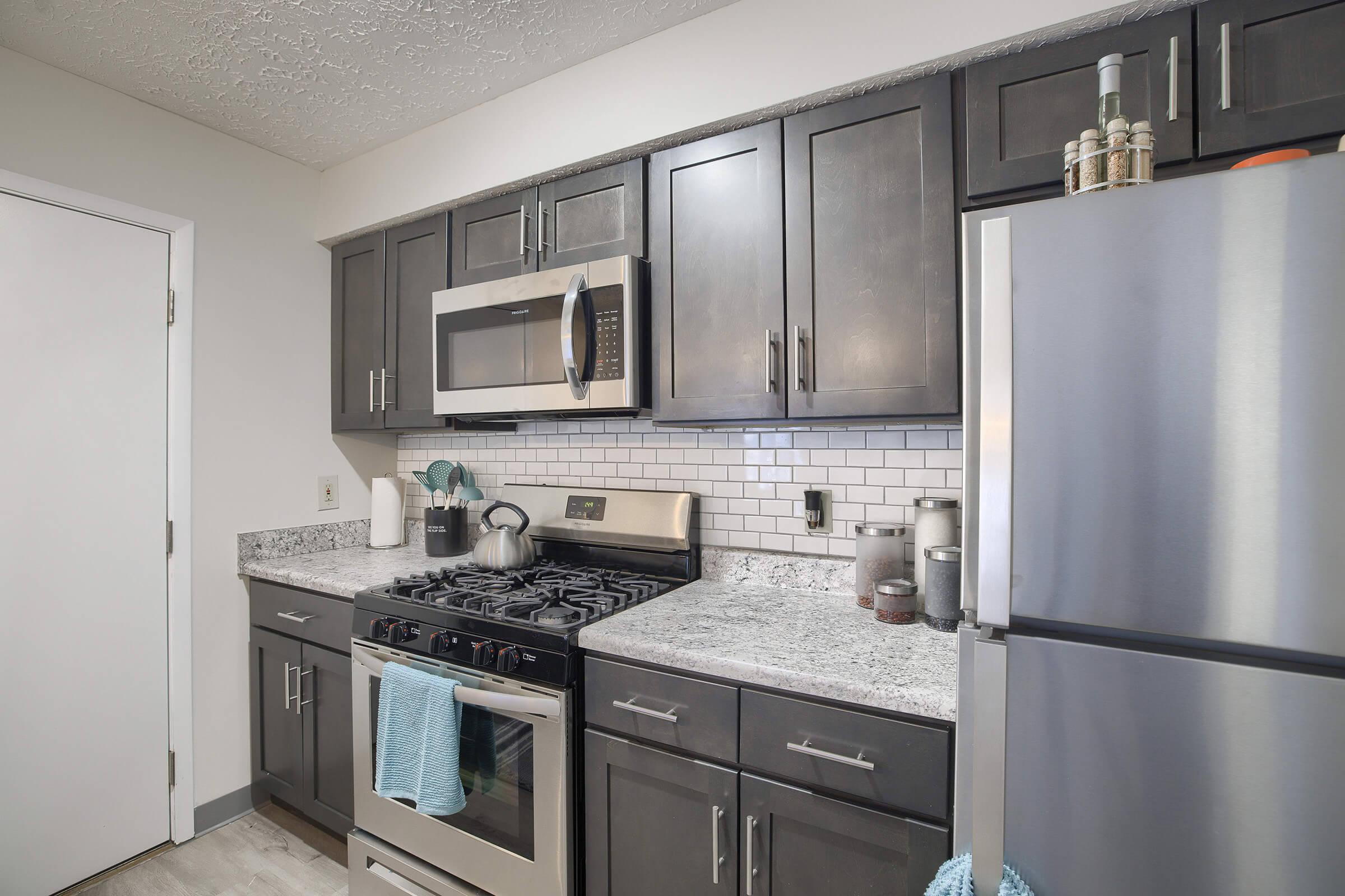 A modern kitchen featuring dark wooden cabinets, a stainless steel refrigerator, and a range with an oven. The countertops are made of gray speckled granite, and a white tile backsplash is present. Small appliances and kitchen utensils are neatly arranged on the counter, creating a clean and organized space.