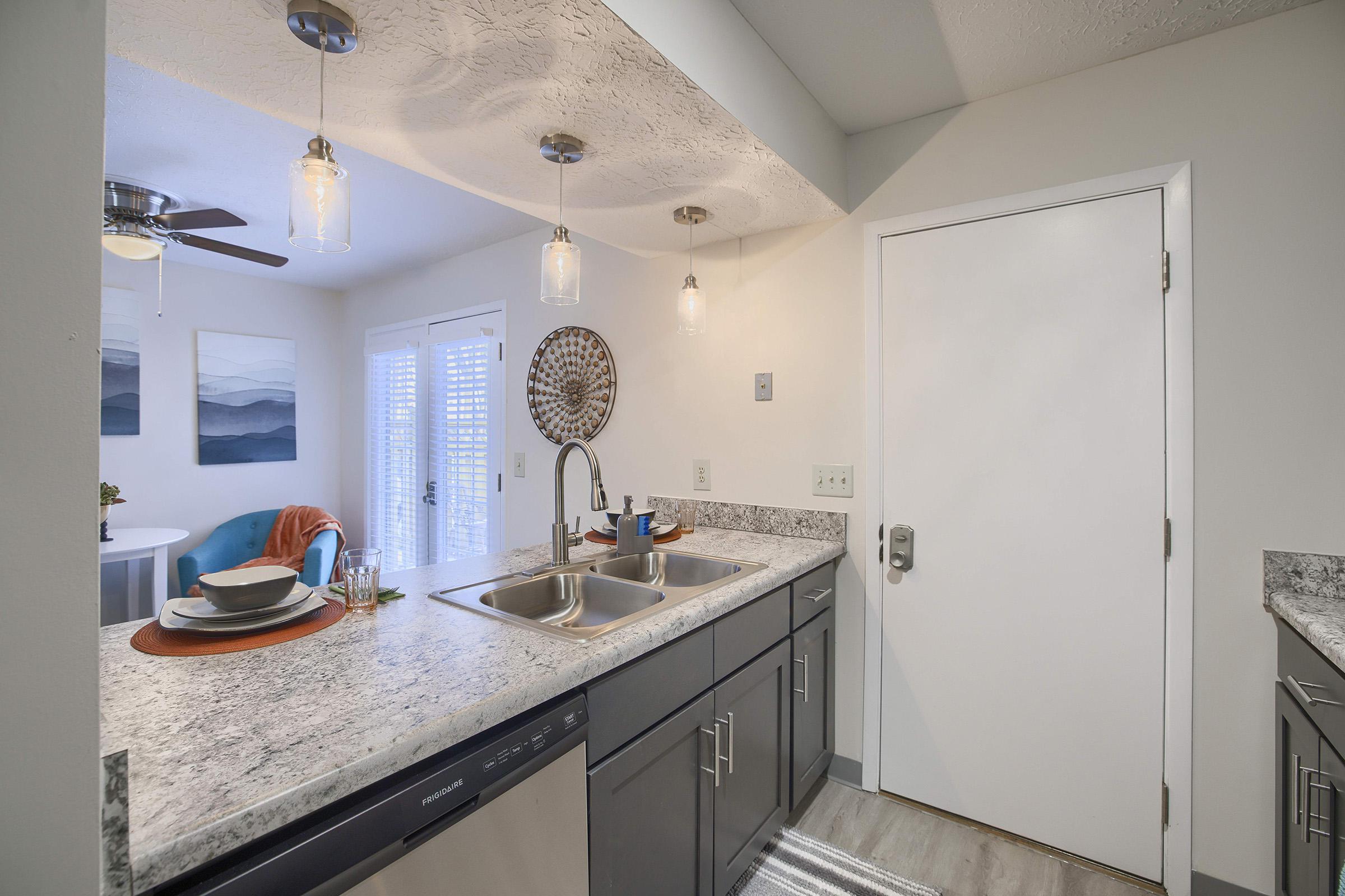 A modern kitchen area featuring a double sink with a countertop, gray cabinets, and pendant lighting. There is a door leading outside, and a small dining space with a blue chair and table in the background. Decor items include a round wall hanging and some dishes on the counter. Bright and inviting atmosphere.