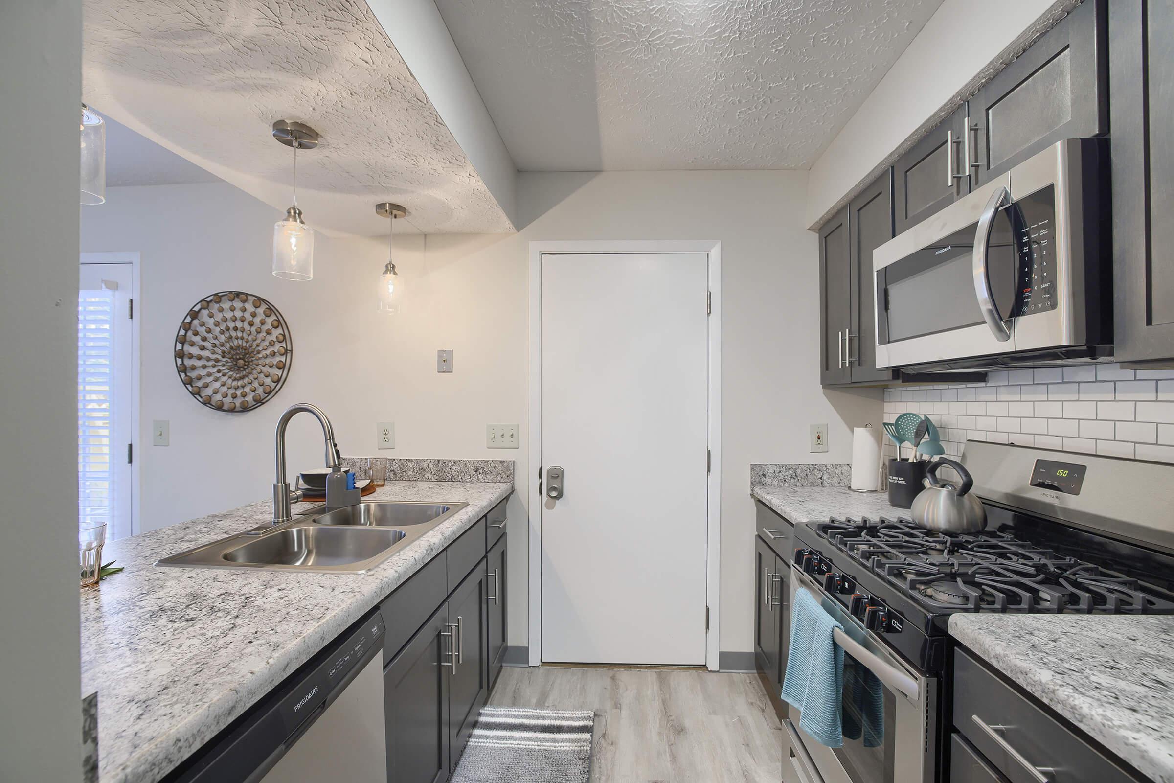 A modern kitchen featuring gray cabinets, a granite countertop, and stainless steel appliances. The space includes a double sink, a gas stove, and a microwave. Natural light enters through a door, and there are decorative elements such as a wall clock and a small plant. The flooring is light-colored and laminate.