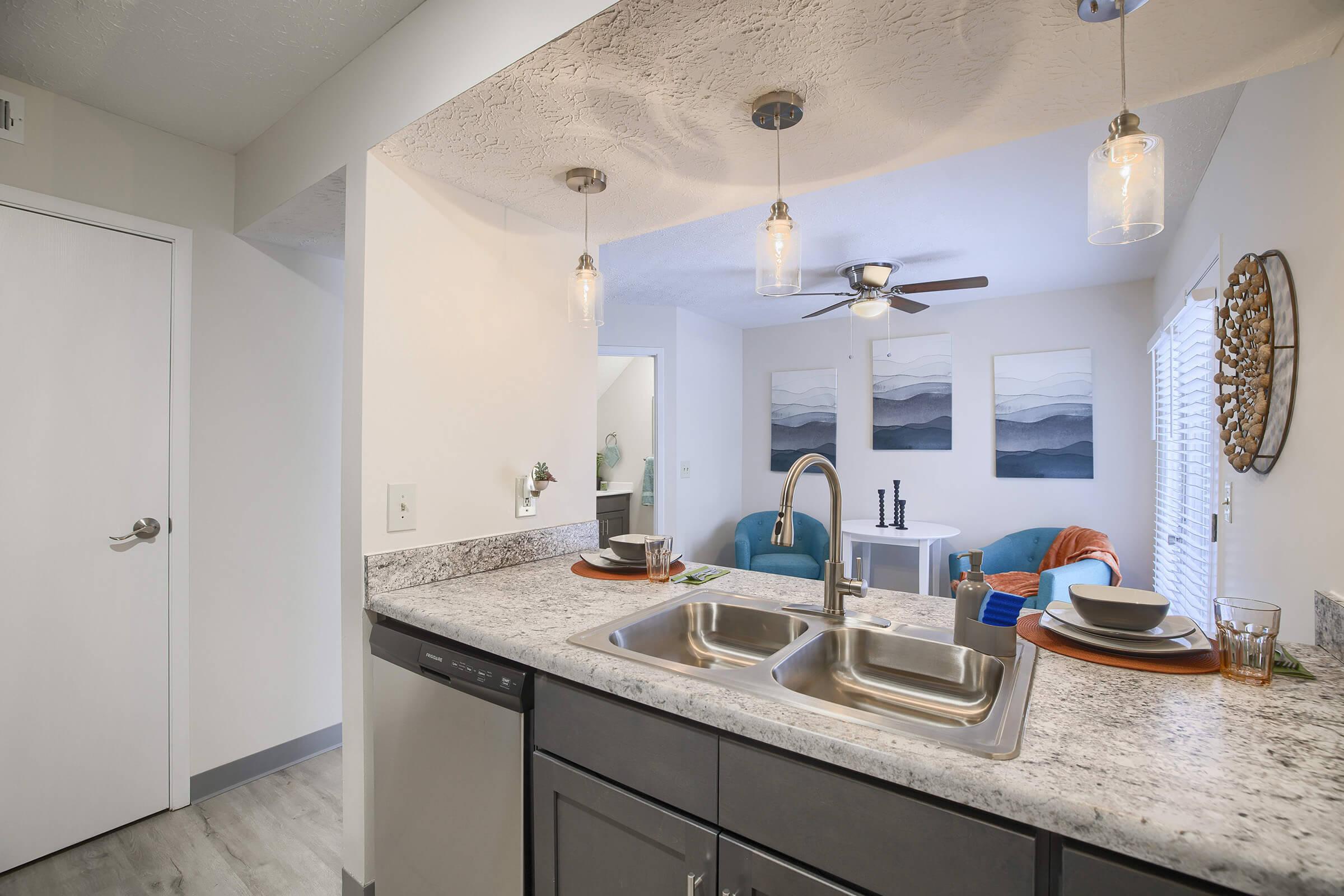 A modern kitchen featuring a stainless steel sink with two basins, a dishwasher, and granite countertops. Above, three pendant lights illuminate the space. In the background, a cozy living area with a ceiling fan, two blue chairs, and decorative wall art can be seen, creating a welcoming atmosphere.